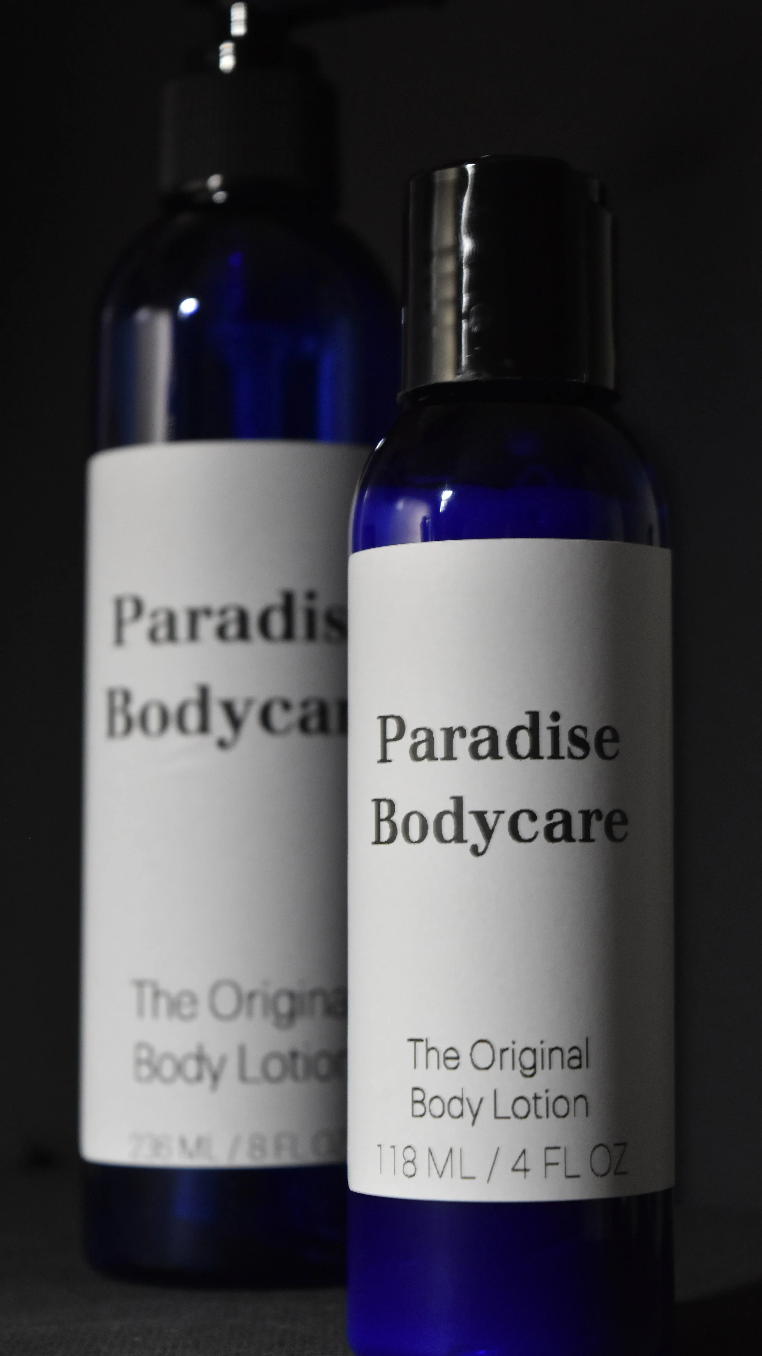 Two blue bottles of Paradise Bodycare lotion with black caps and white labels. One bottle is in the foreground, slightly to the right, and the other is blurred in the background.