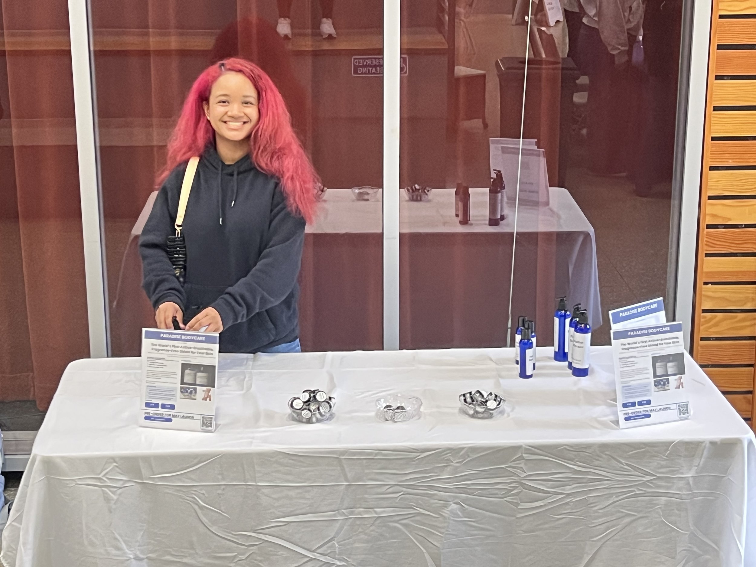 A woman with pink hair smiling at a table with informational pamphlets, small containers, and bottles, behind a glass partition.