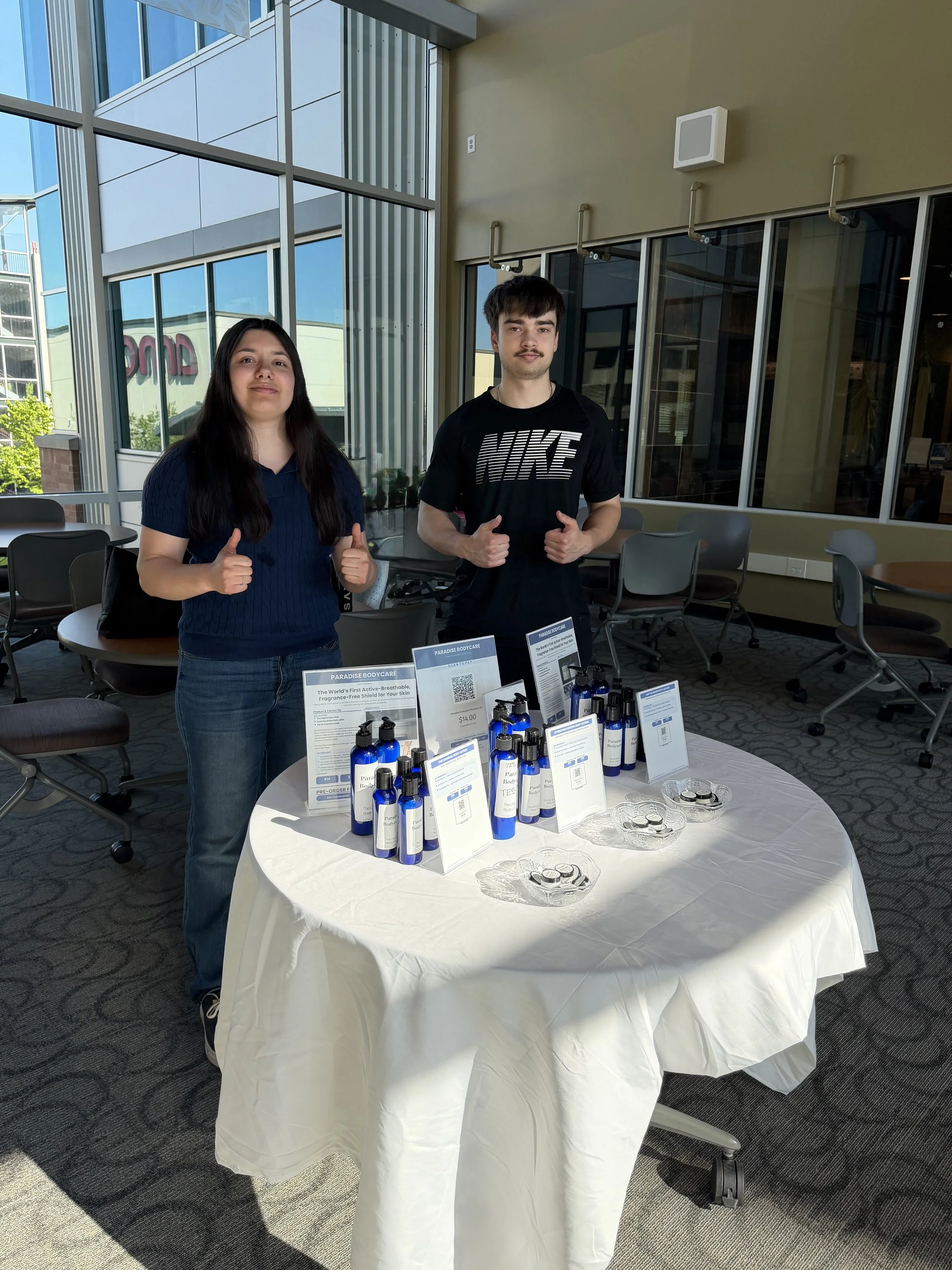 Two young adults standing behind a round table displaying skincare products in bottles and informational cards, inside a modern building with large windows.