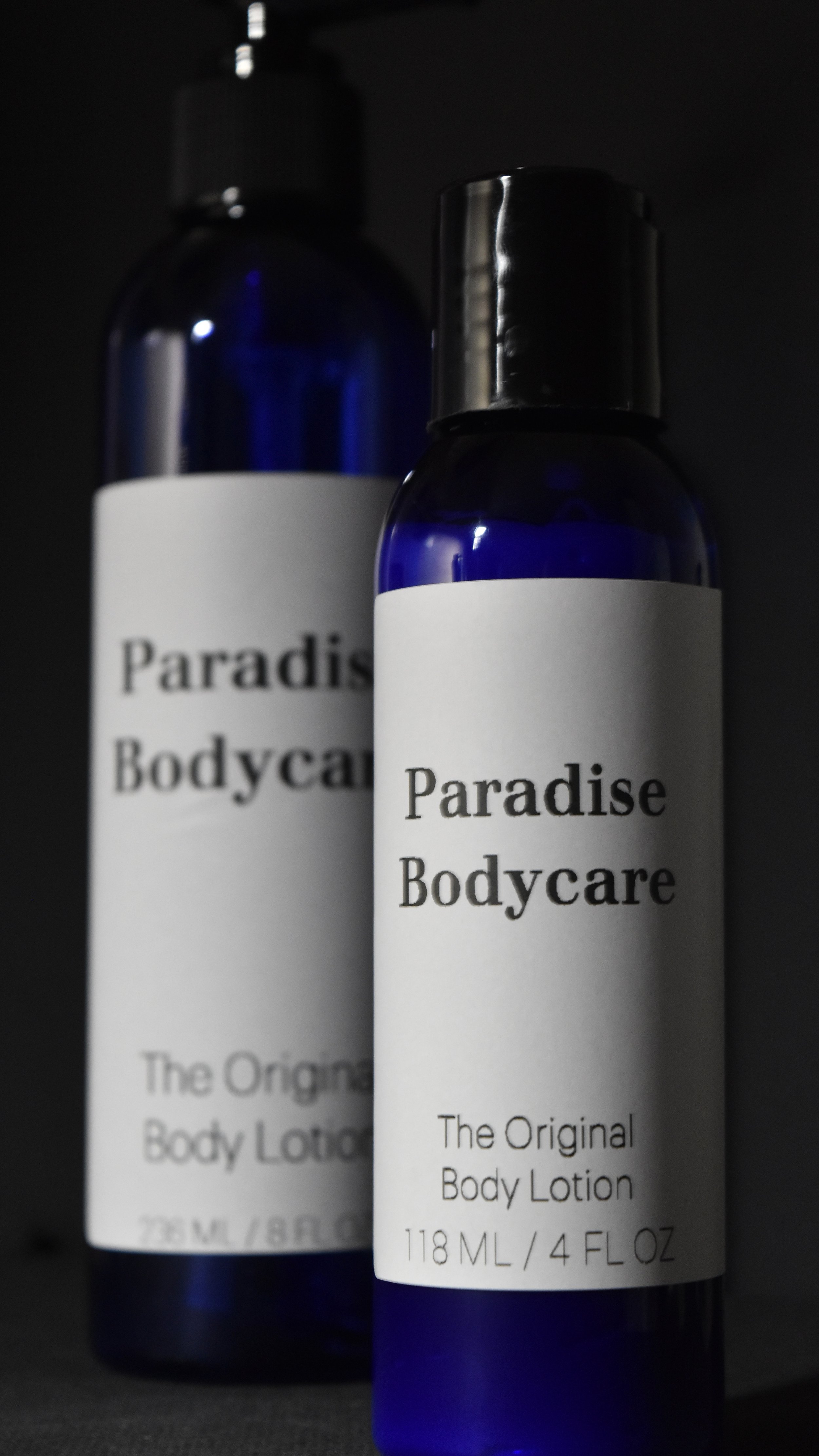 Two blue bottles of Paradise Bodycare body lotion with white labels and black caps, with one bottle in focus and the other slightly blurred in the background.
