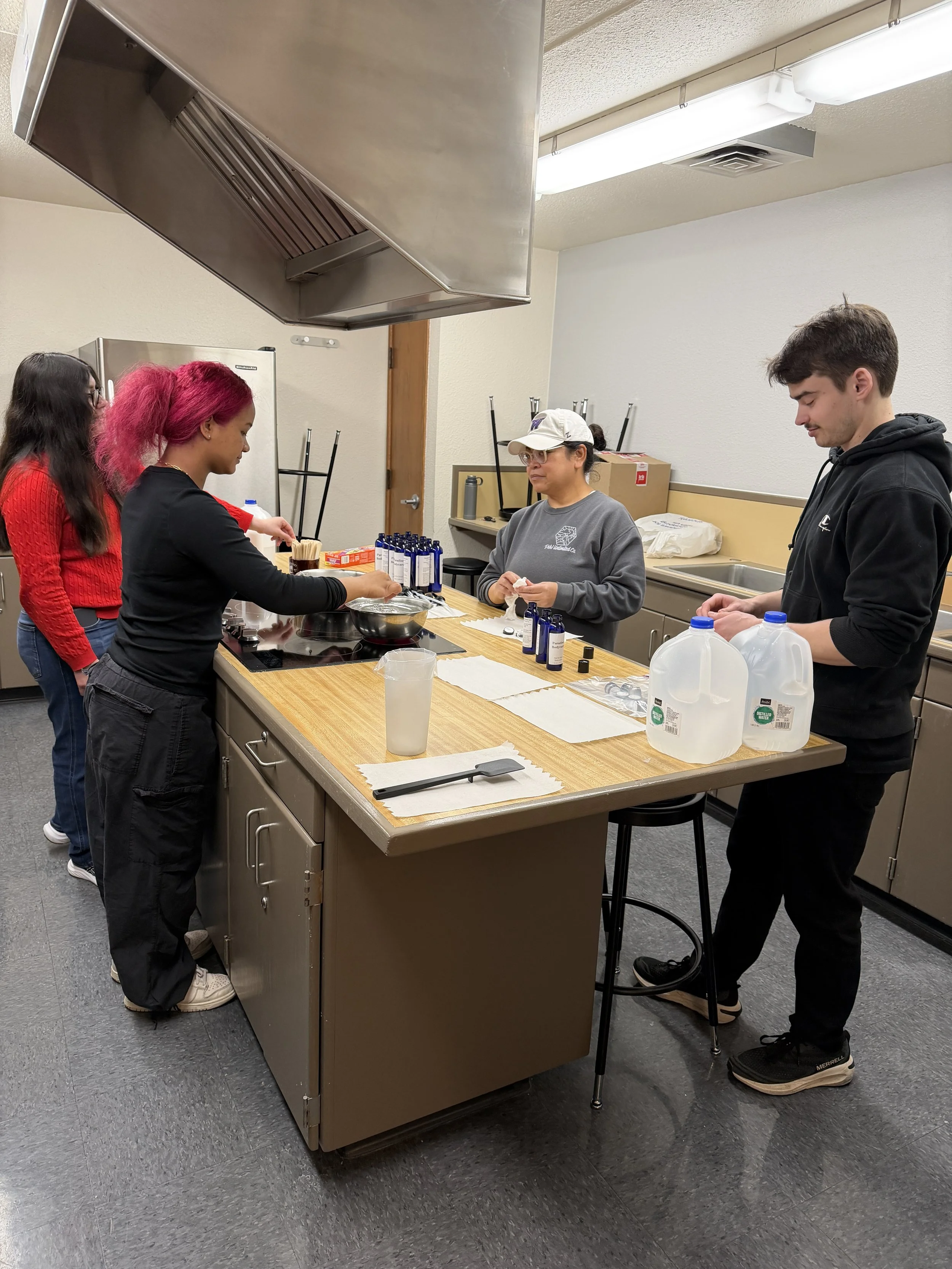 Five people standing around a kitchen counter with bottles of liquid, paper towels, and equipment. One woman in a gray sweatshirt appears to be preparing something while others observe or wait.