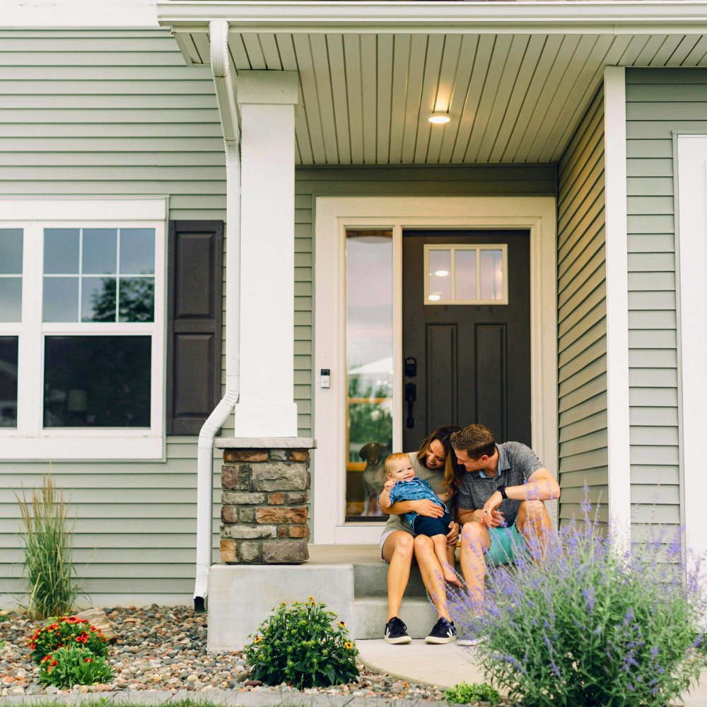 Family sitting on front porch of a house, holding a toddler, with a dog visible through the glass door.