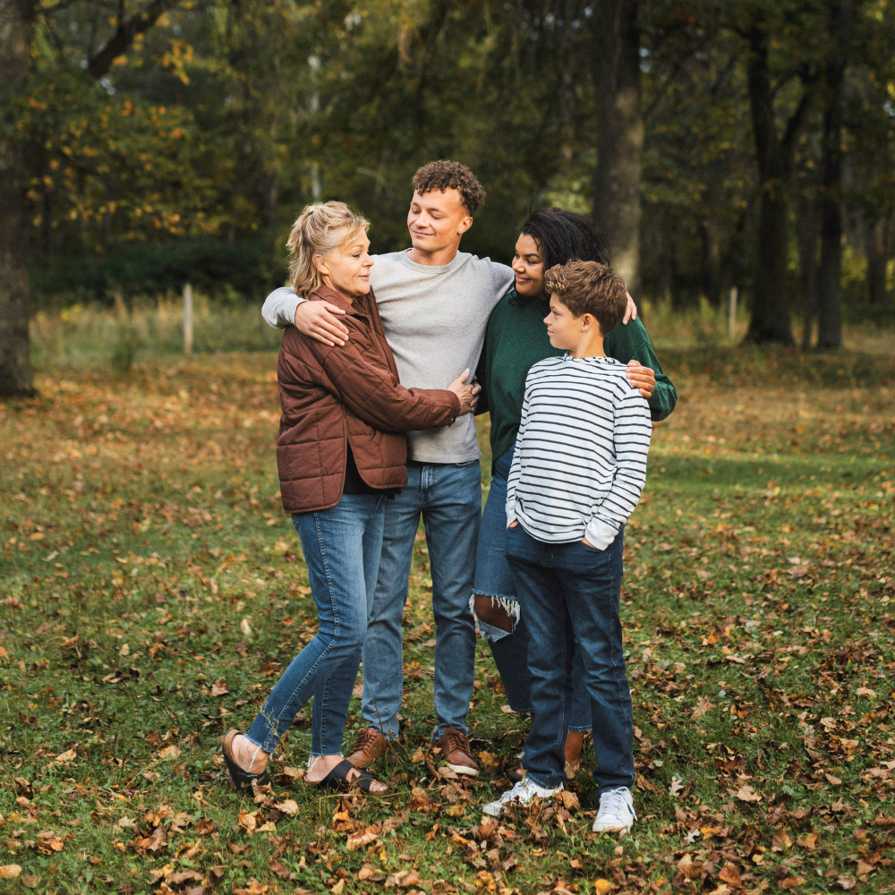 Four people embracing in a park during fall, surrounded by trees with orange and yellow leaves.
