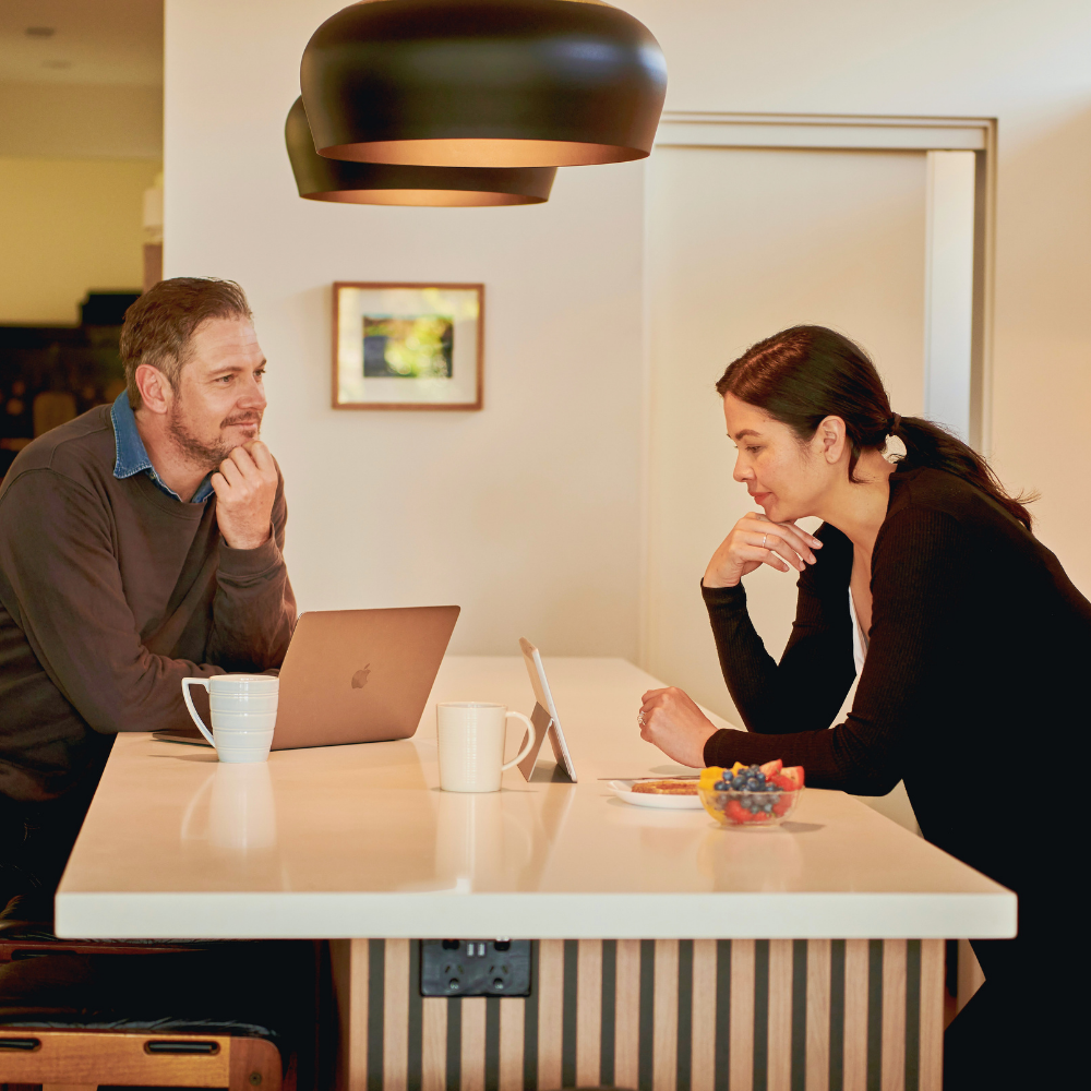 A man and a woman having a serious conversation at a kitchen island, with a laptop, a tablet, cups, and a bowl of berries and fruit on the counter.