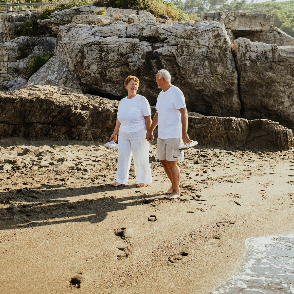 An elderly couple walking hand in hand on a sandy beach with footprints visible in the sand, large rocks in the background, and the ocean waves washing ashore.