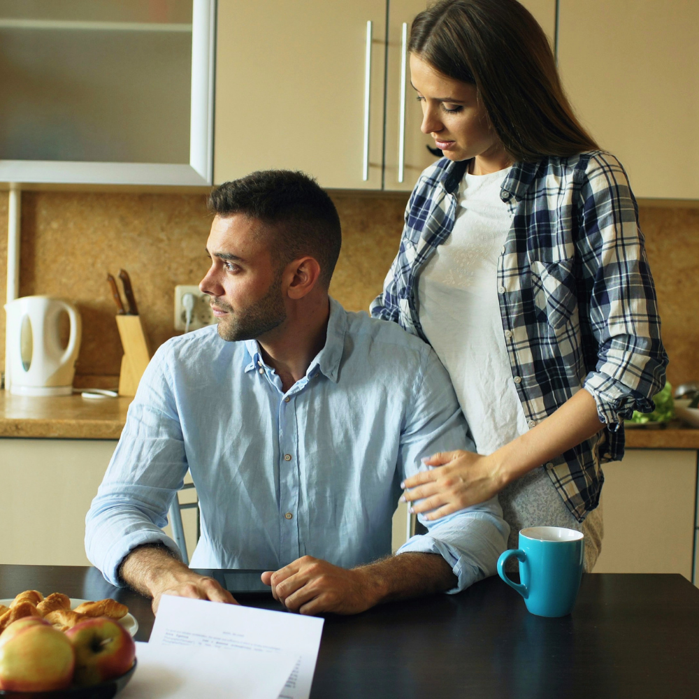 A woman stands beside a seated man in a kitchen, gently touching his arm. There are apples and croissants on the table in front of them, and a blue mug next to the woman.