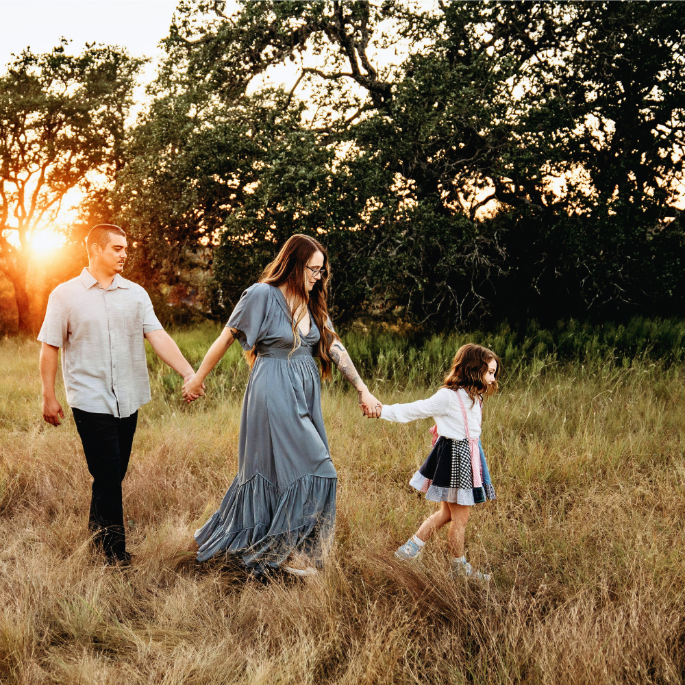 A family of four holding hands and walking through a grassy field during sunset, with large trees in the background.