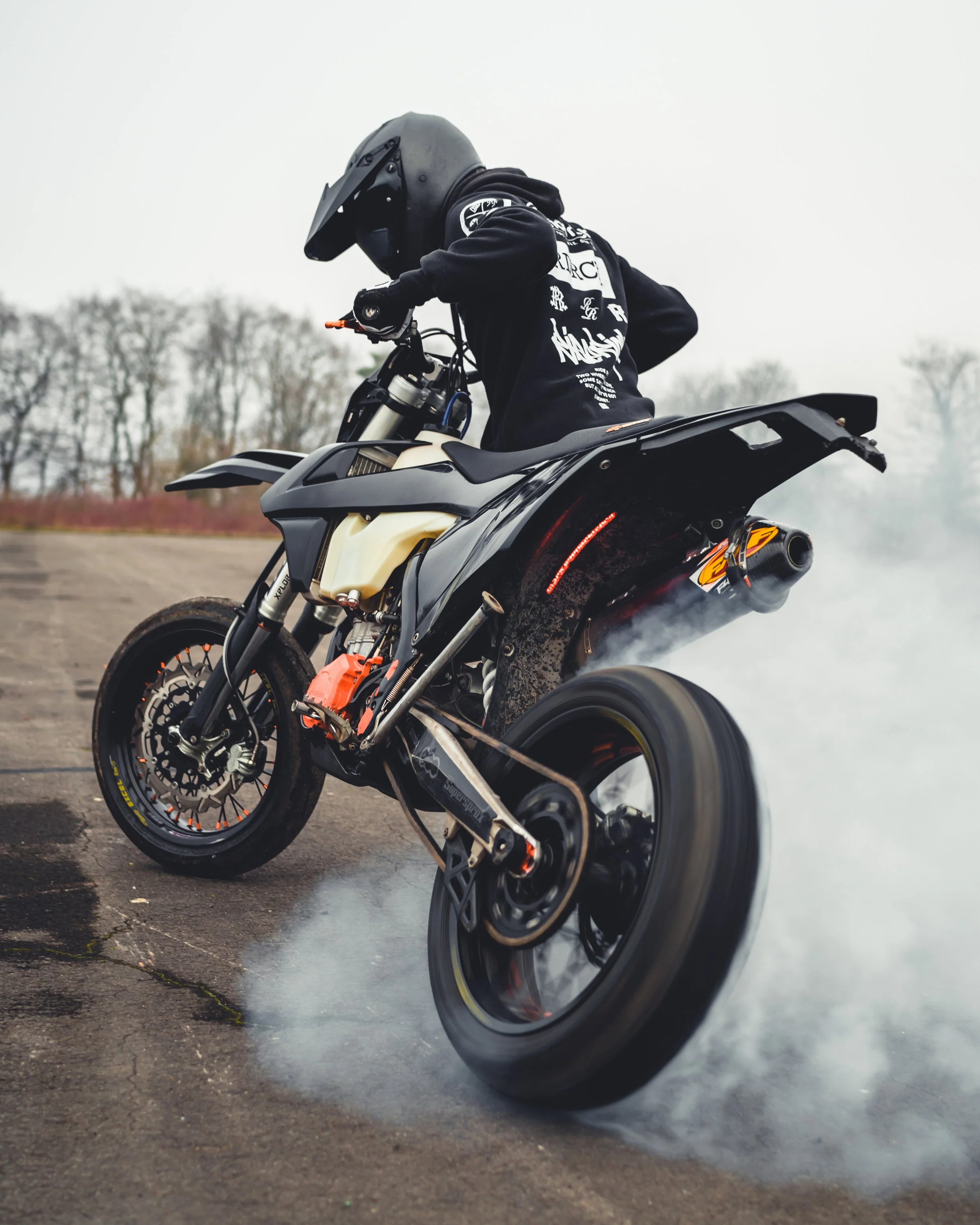 A person performing a burnout on a black dirt bike, wearing a black helmet and black hoodie, on an open paved area with smoke and leafless trees in the background.