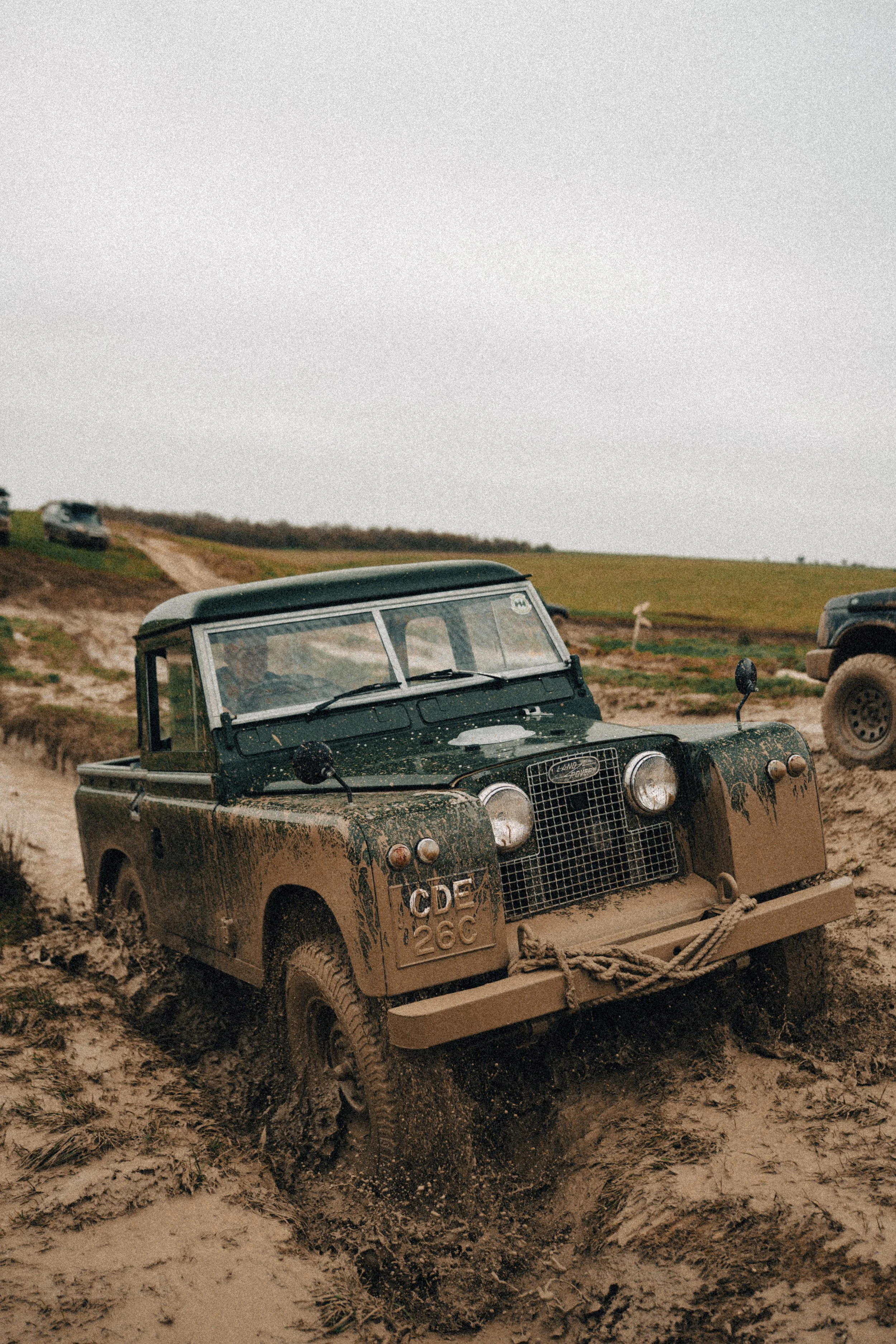 A muddy Land Rover Defender driving through a wet, muddy off-road terrain on a cloudy day.
