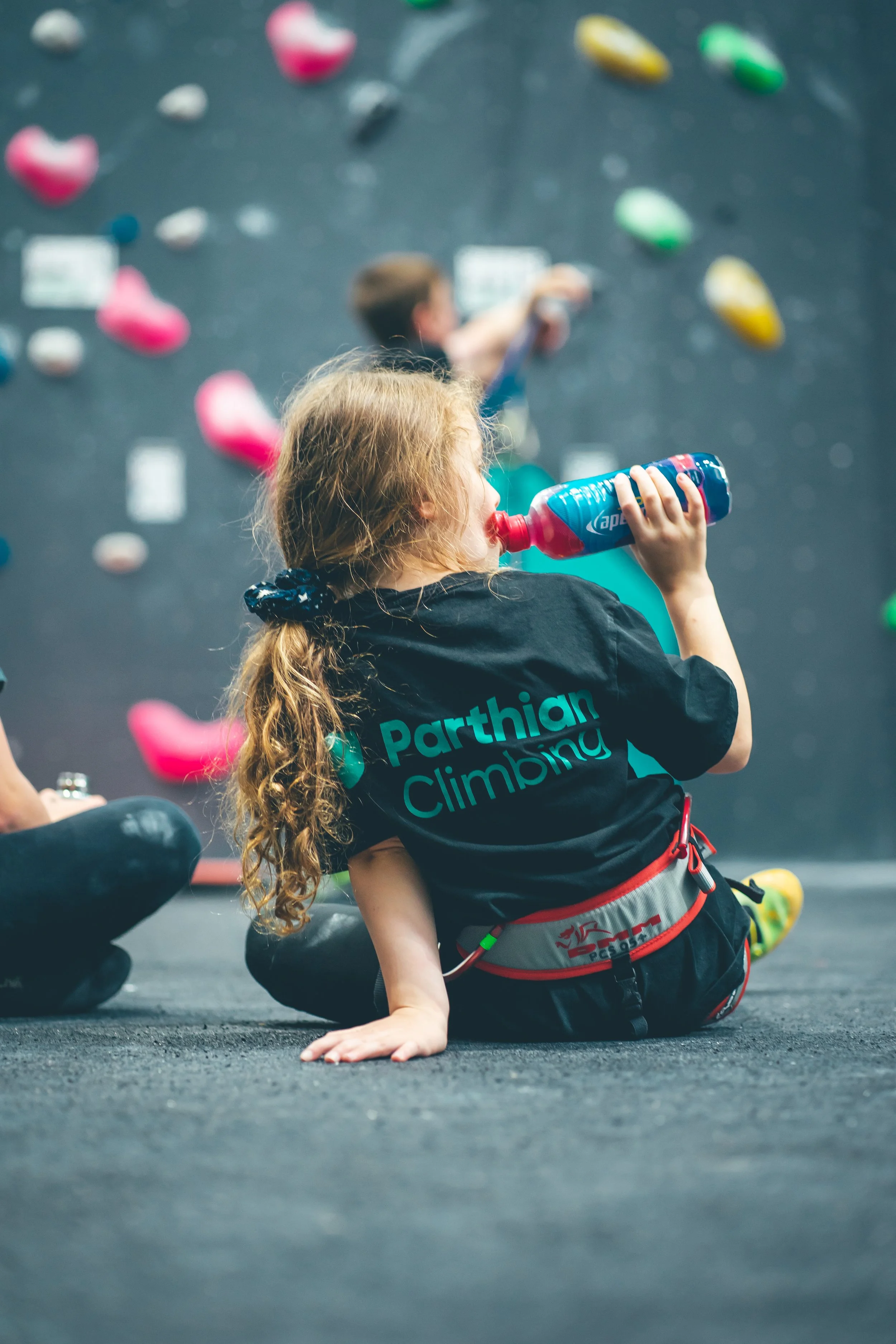 A young girl with curly hair drinking water while sitting on the floor of an indoor climbing gym, with a climbing wall and colorful holds in the background.