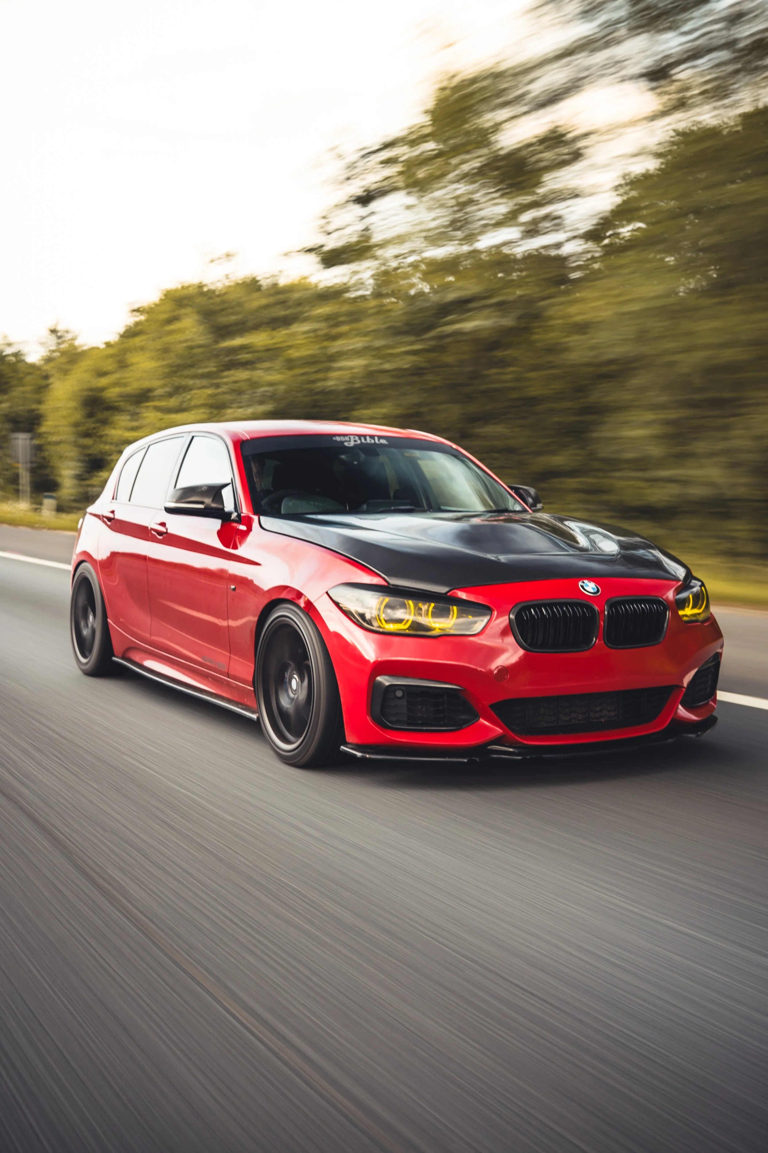 A red BMW car driving fast on an open highway with a blurred green wooded background.