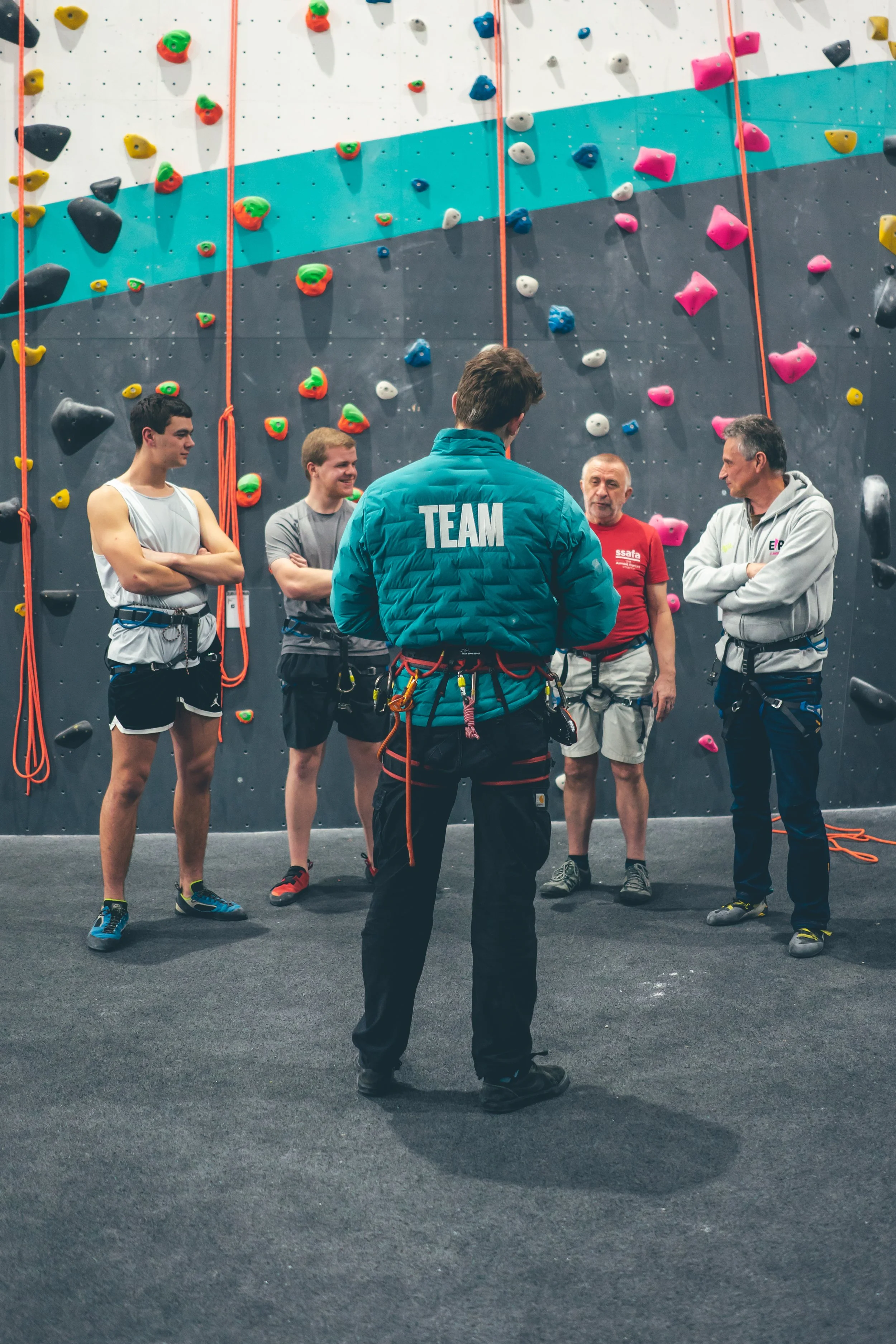 Group of five people with climbing gear, standing in front of an indoor rock climbing wall with colorful holds, having a discussion.
