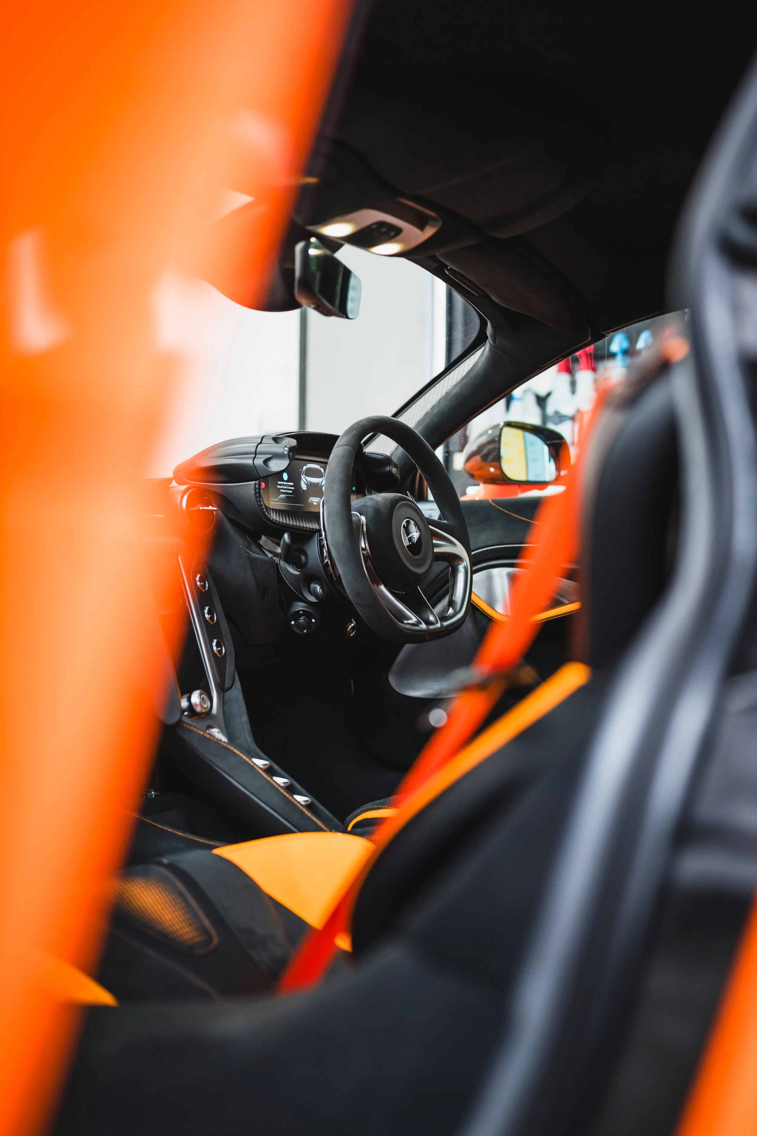 Interior of a luxury sports car with a black steering wheel, orange accents, and a digital dashboard, viewed through the gaps of the car's orange and black interior panels.