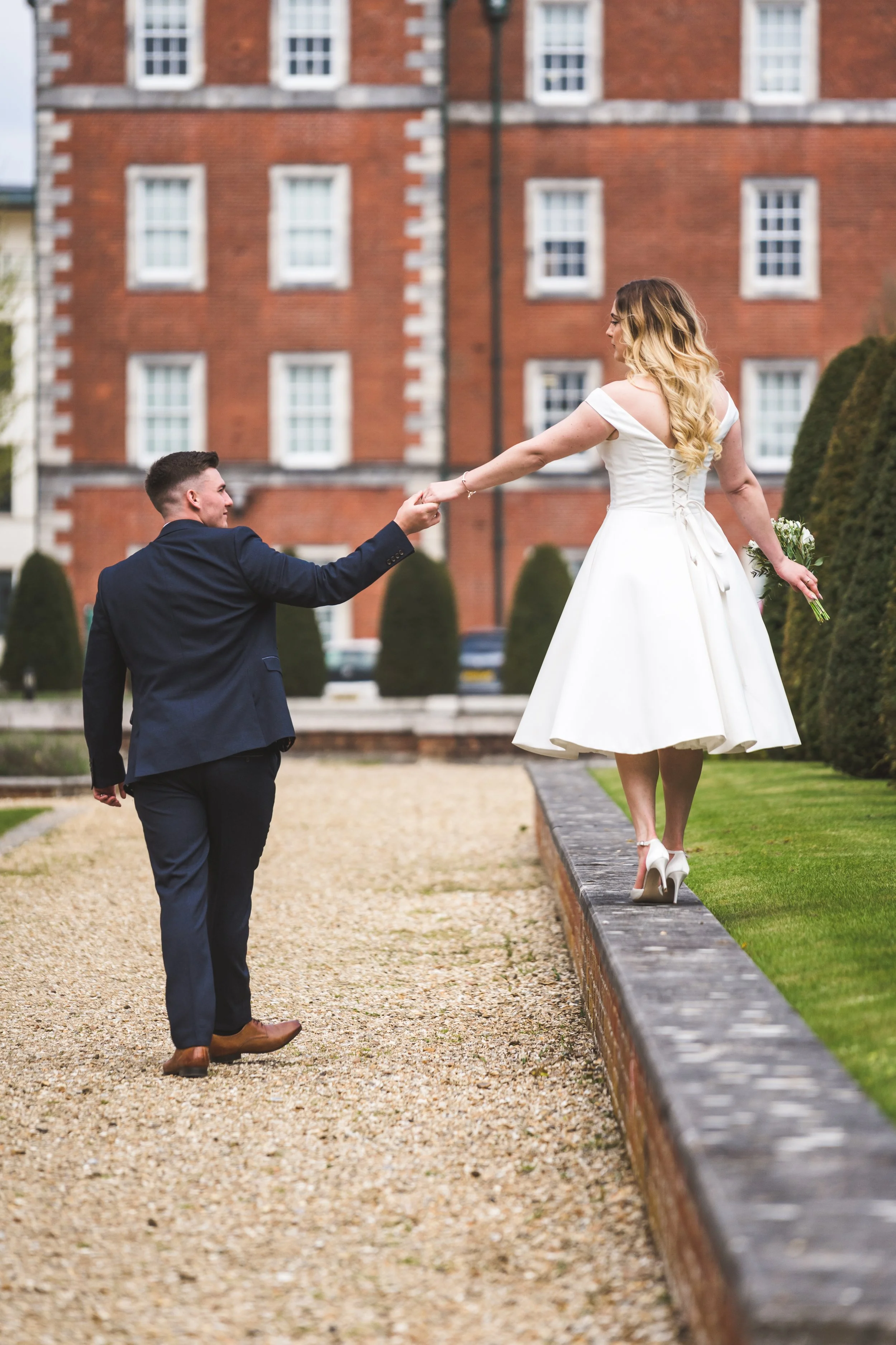 A groom helping a bride walk along a low wall outside a red brick building with white window frames, during a wedding photoshoot.