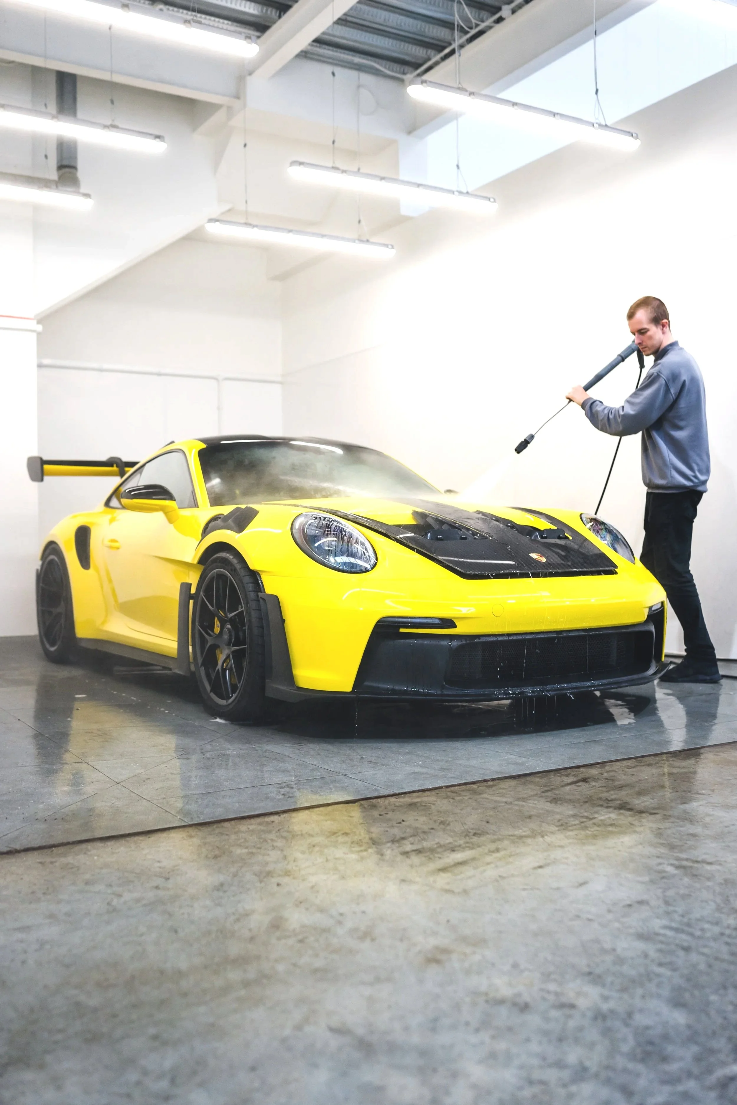 A man washing a yellow sports car with a pressure washer in a car wash facility.