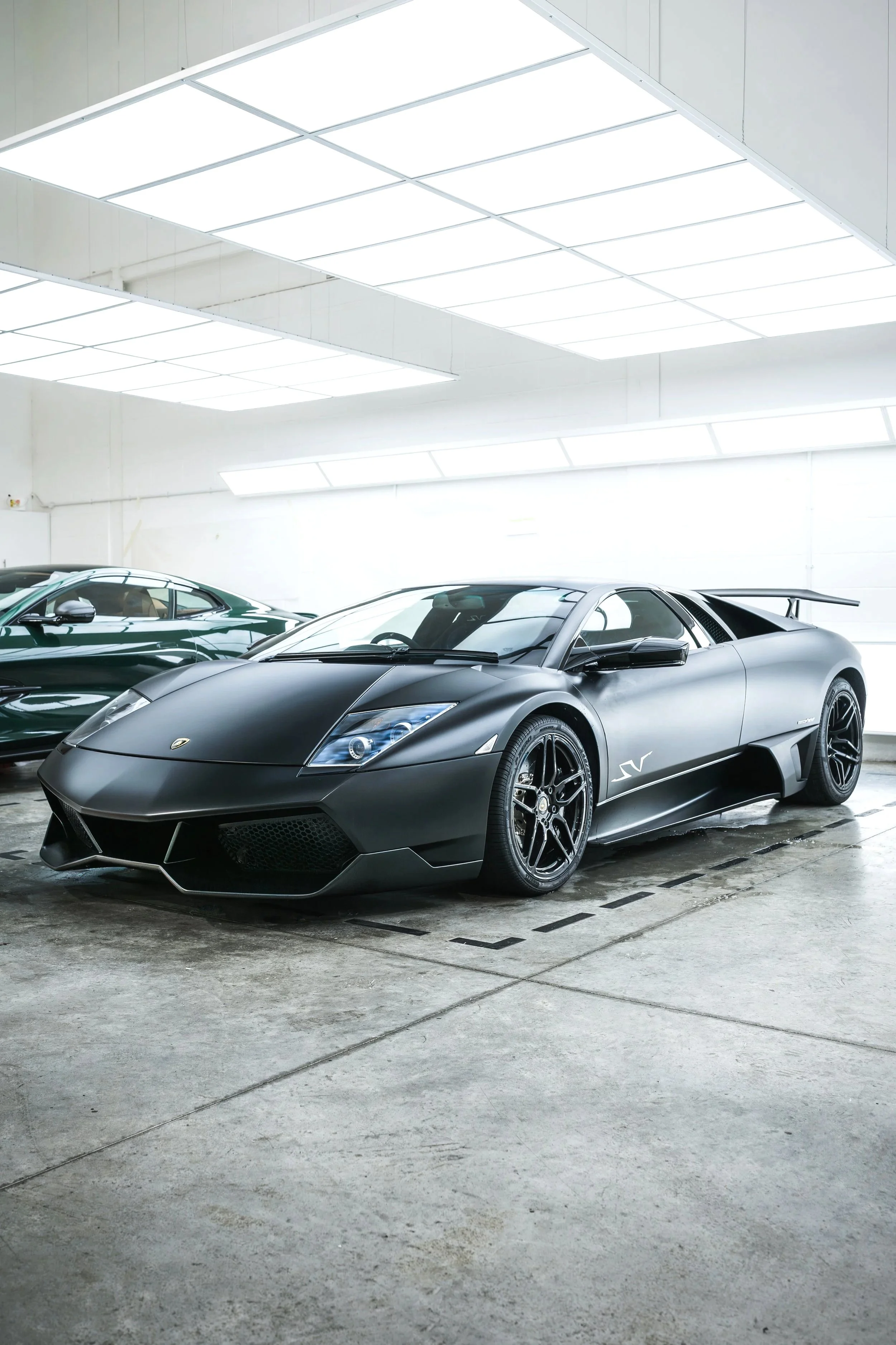 Black luxury sports car, Lamborghini, parked indoors next to a green car in a well-lit garage.