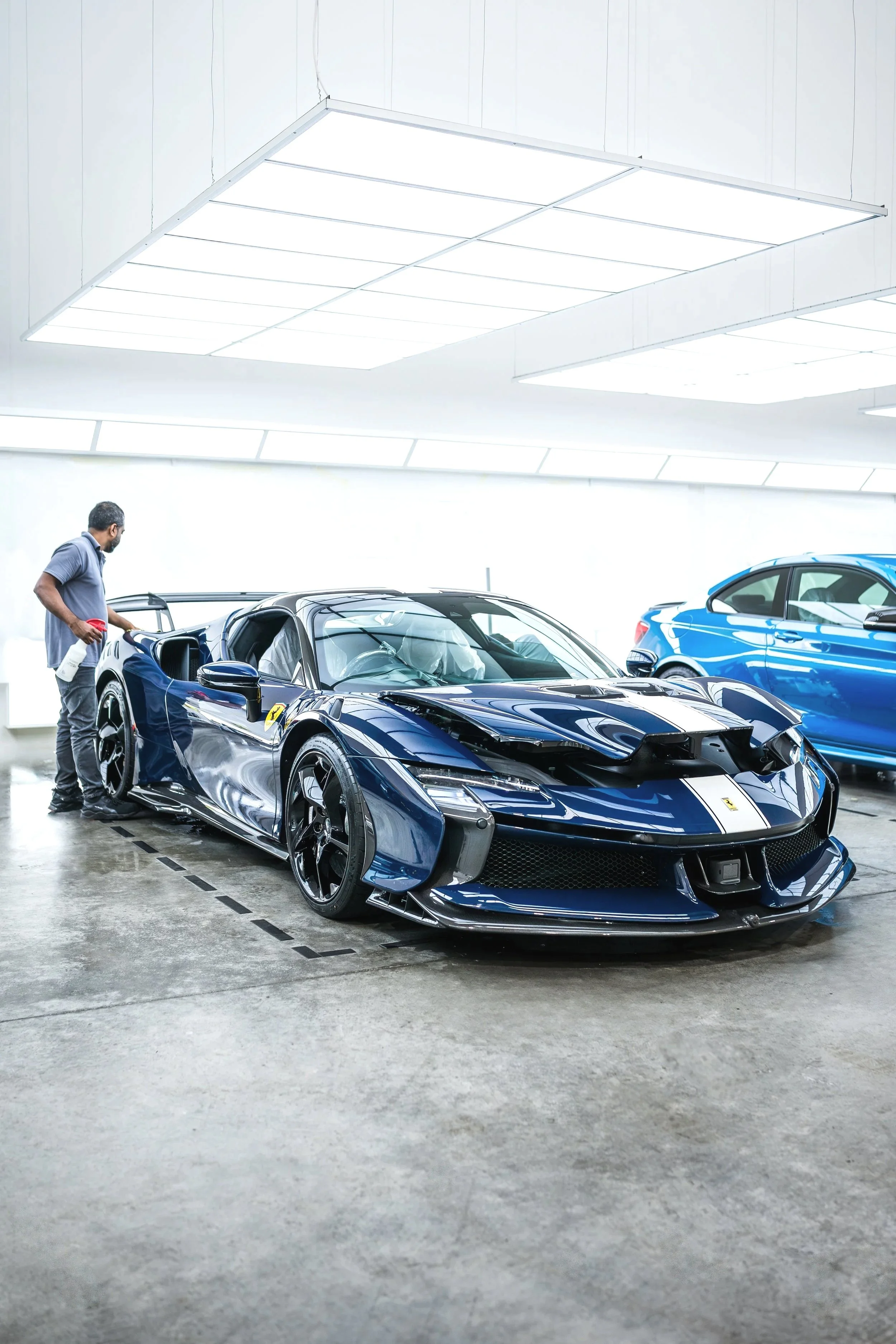 A man cleaning a blue Ferrari sports car inside a showroom, with another blue vehicle in the background.