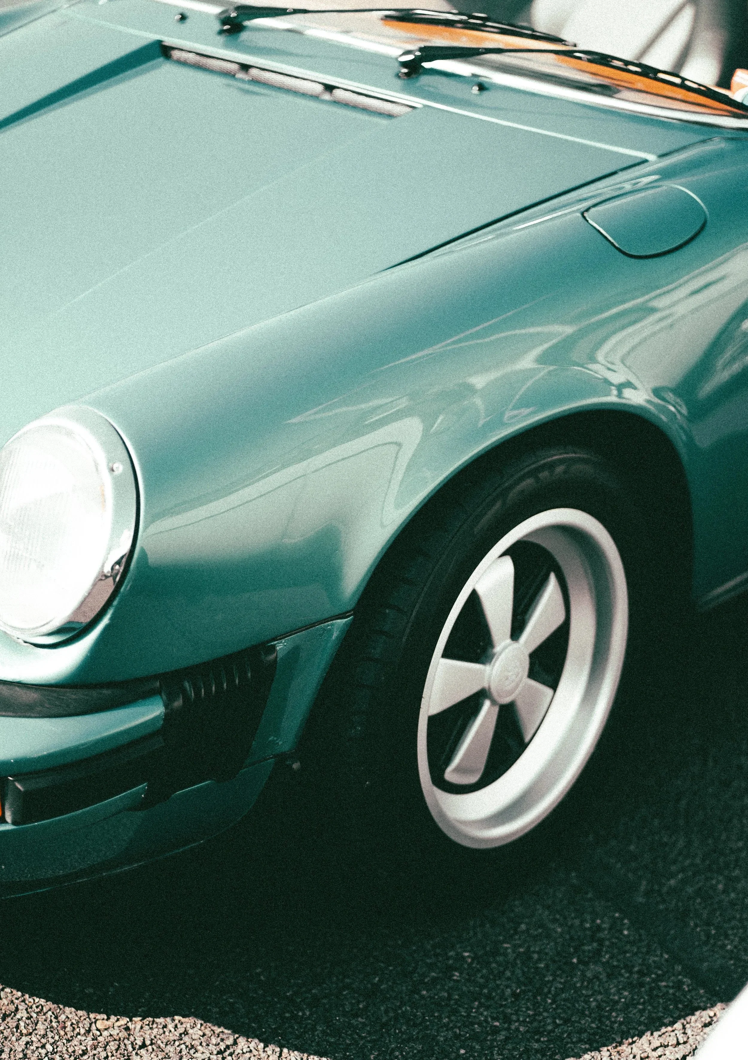 Close-up of a vintage teal sports car showing front left headlight, wheel, and part of the hood with a chrome windshield wiper.