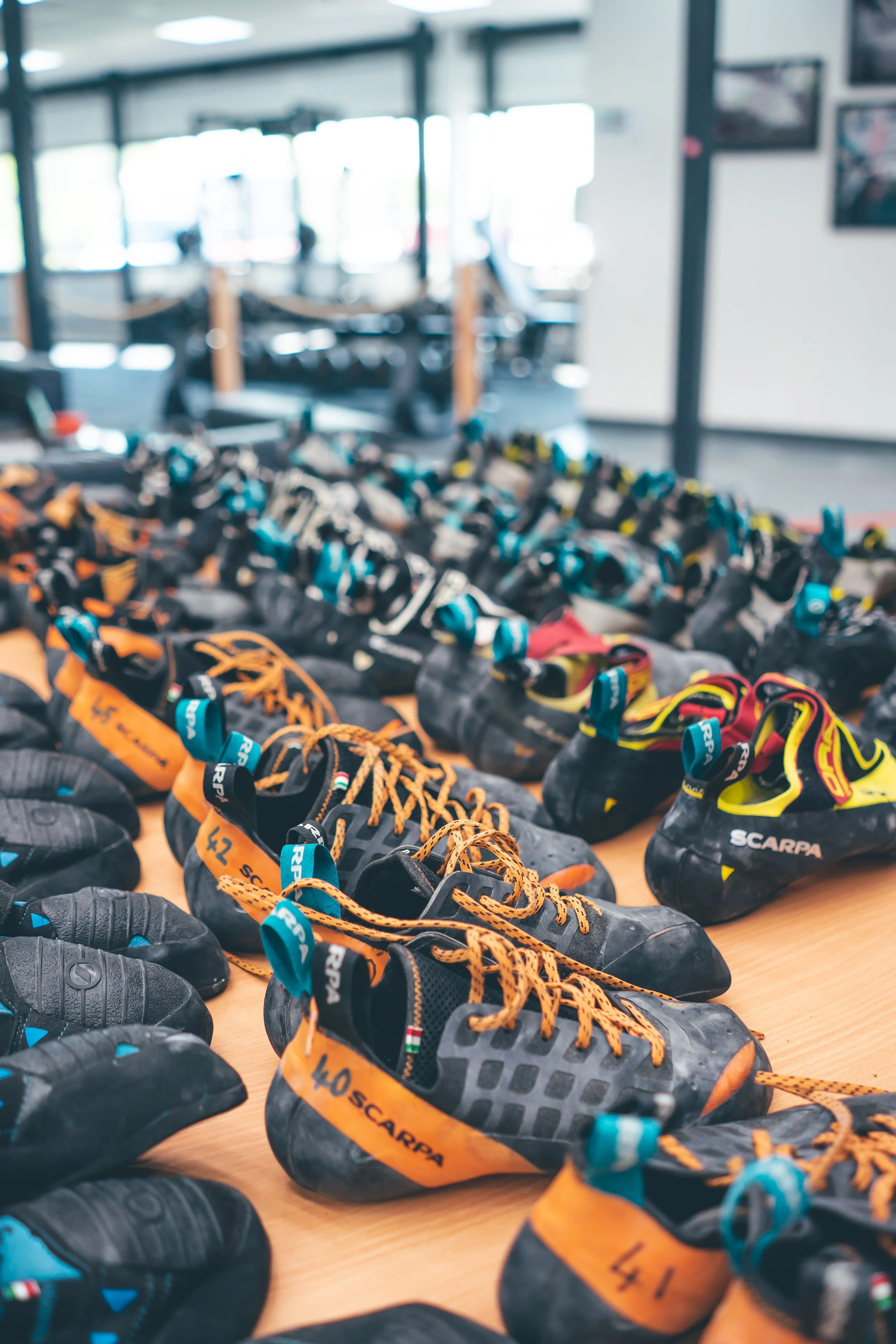 Multiple pairs of climbing shoes with labels on a wooden table inside a climbing gym.