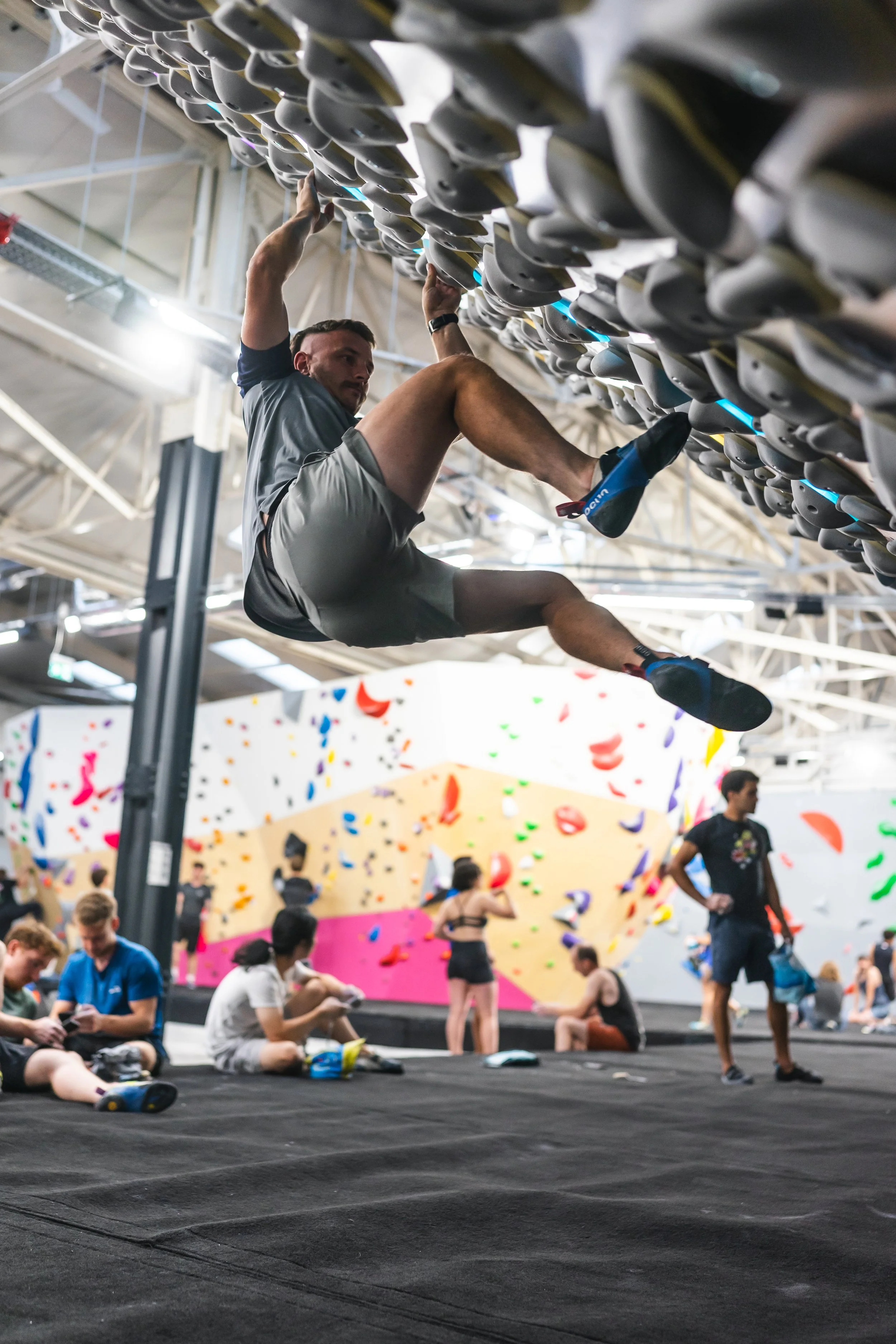 A man is mid-air on an indoor rock climbing wall, hanging from holds above him. Other climbers and spectators are visible in the background, with colorful climbing walls behind them.