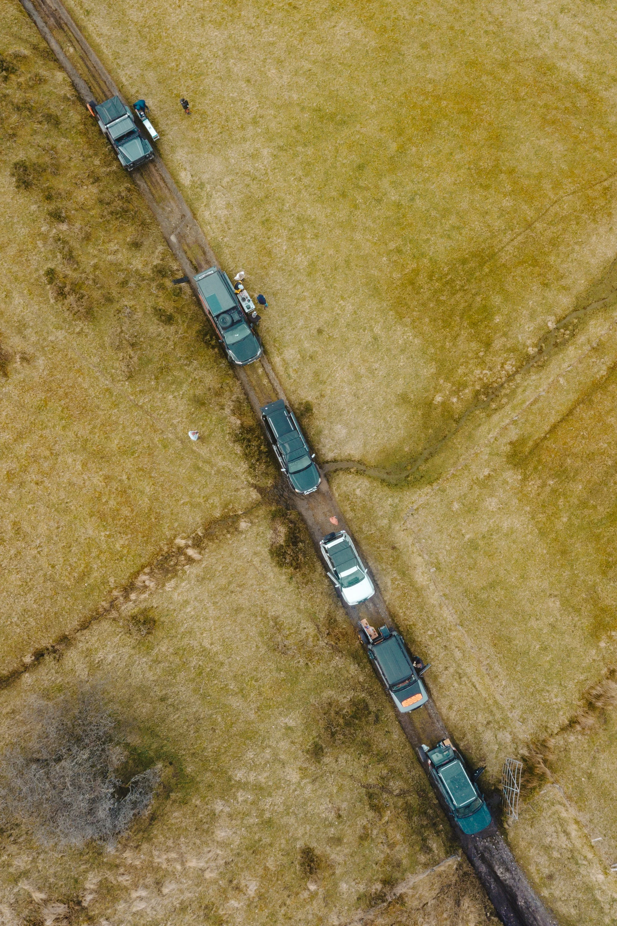 Aerial view of a narrow dirt road with several parked trucks and people standing nearby, surrounded by fields.
