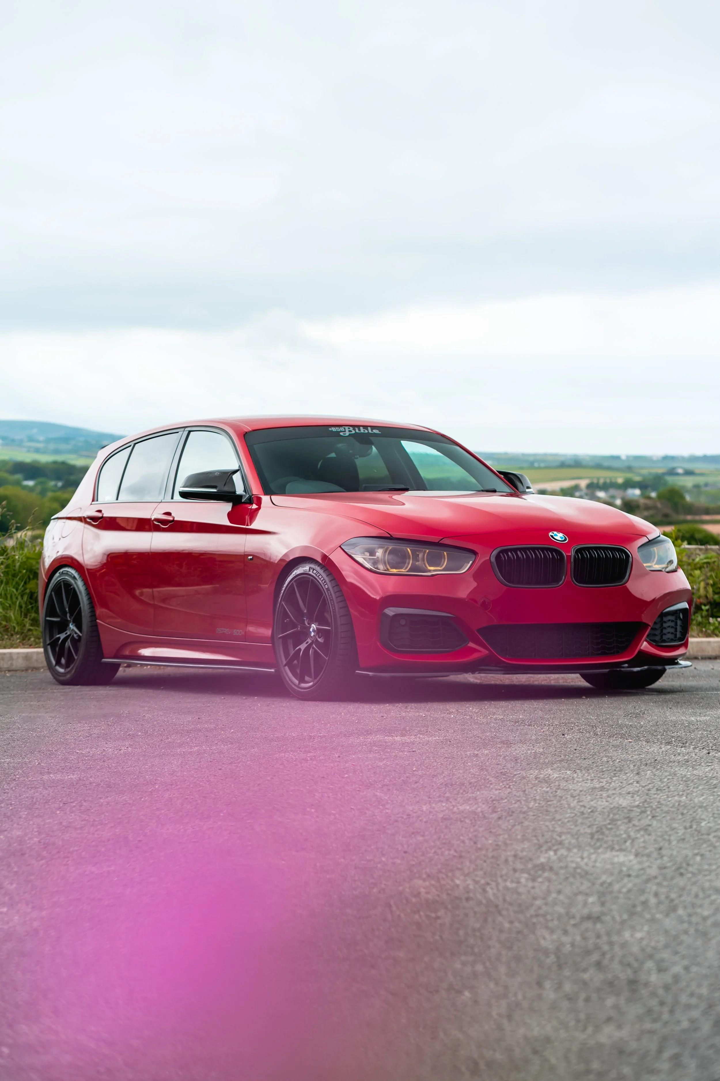 Red BMW hatchback parked on the road with mountainous landscape in the background.
