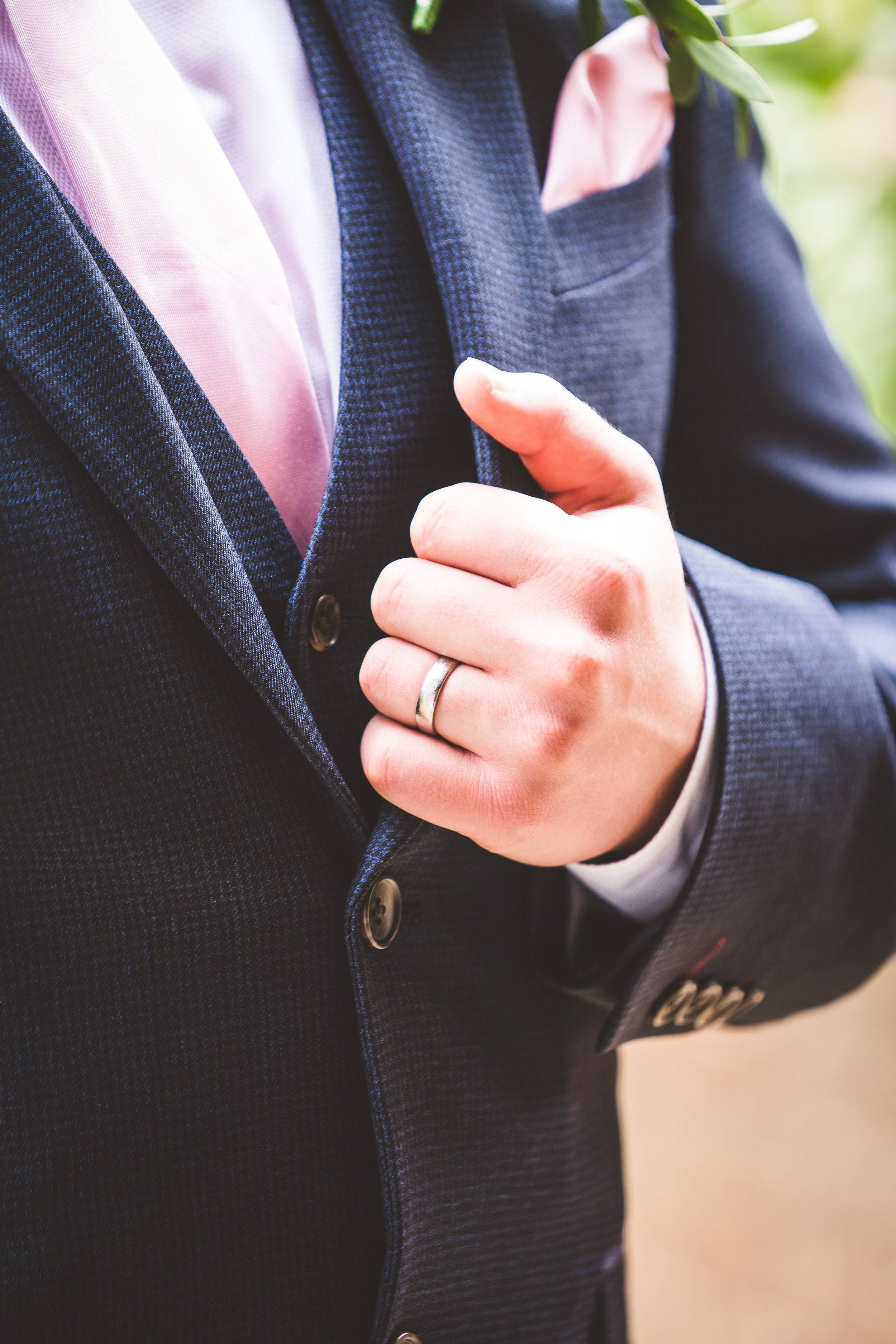 Close-up of a man in a dark suit with a pink pocket square, wearing a wedding band, holding the lapel of his suit jacket.