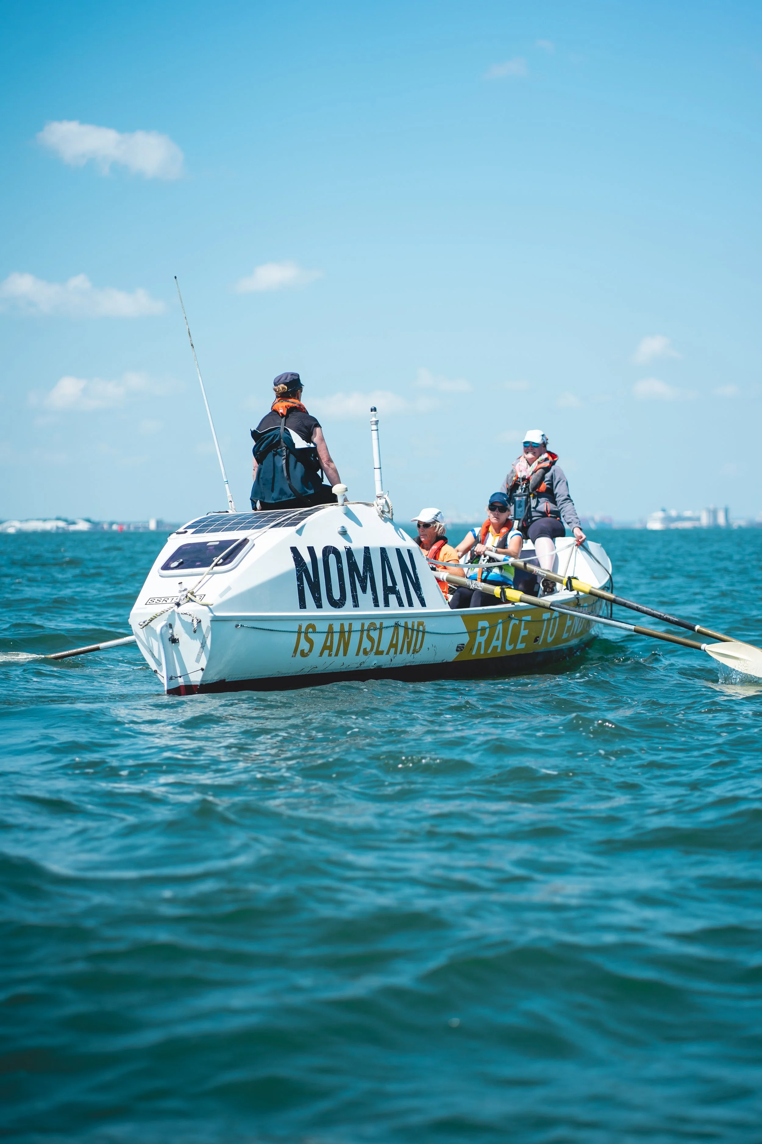 A team of rowers participating in a sailing race on a boat named 'NOMAN,' with a clear blue sky and calm water in the background.