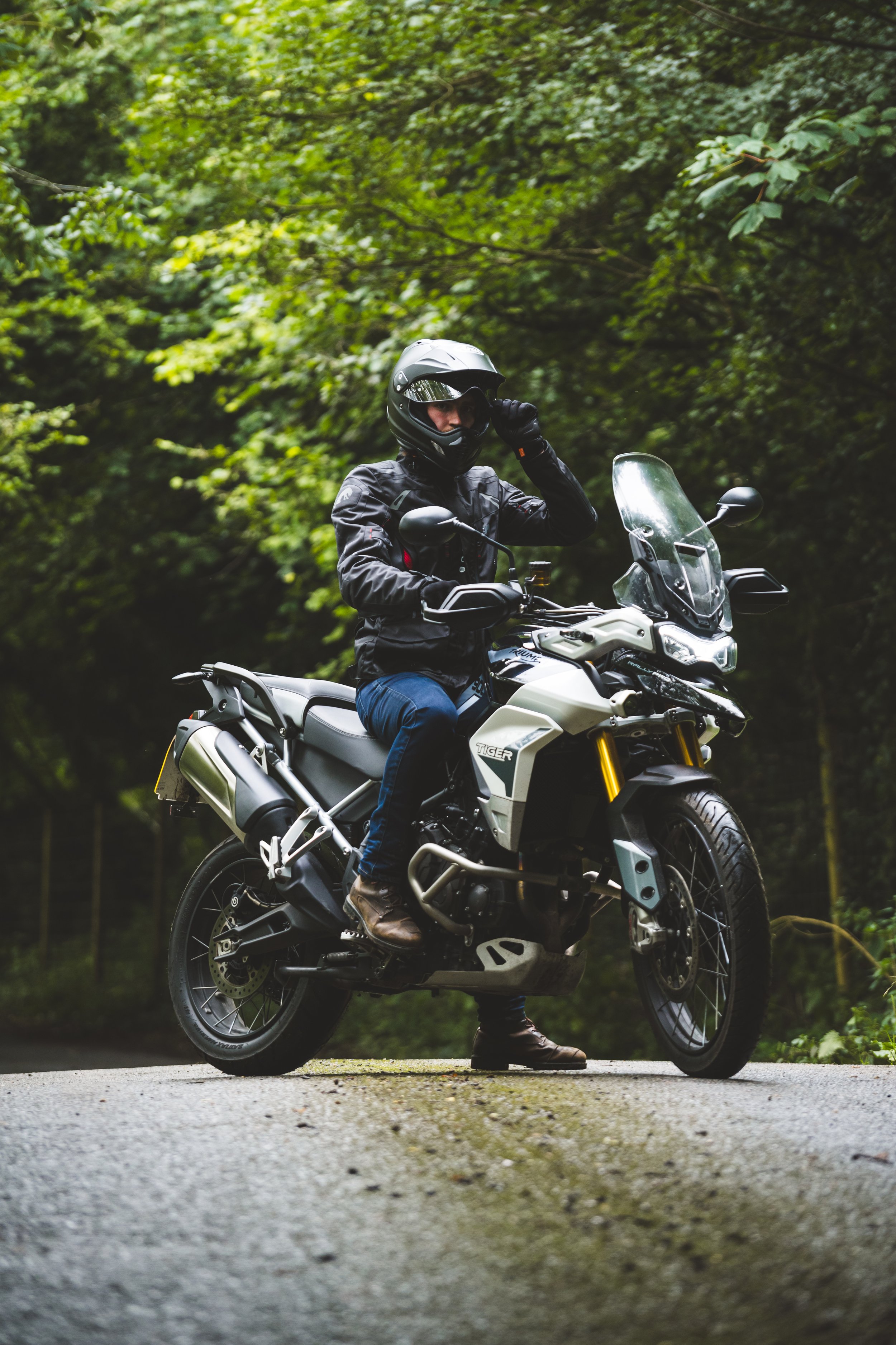 A person wearing a black helmet, black jacket, and brown boots adjusts their helmet while sitting on a white adventure motorcycle on a forested road.