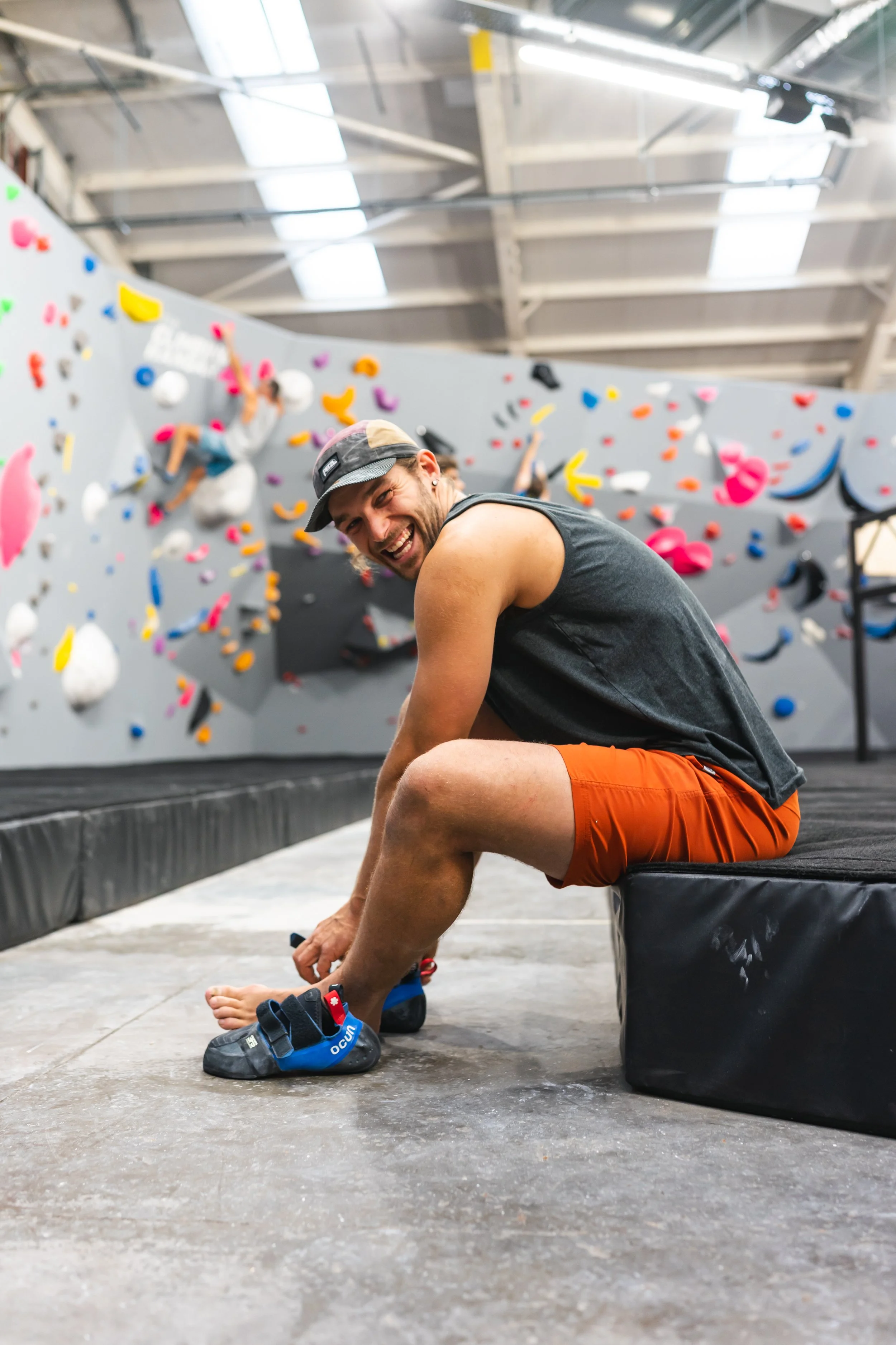 A man in a black sleeveless shirt, orange shorts, and a cap sitting on a bench at an indoor climbing gym. He is smiling while tying his climbing shoes. The background features a large bouldering wall with colorful holds and another climber visible in