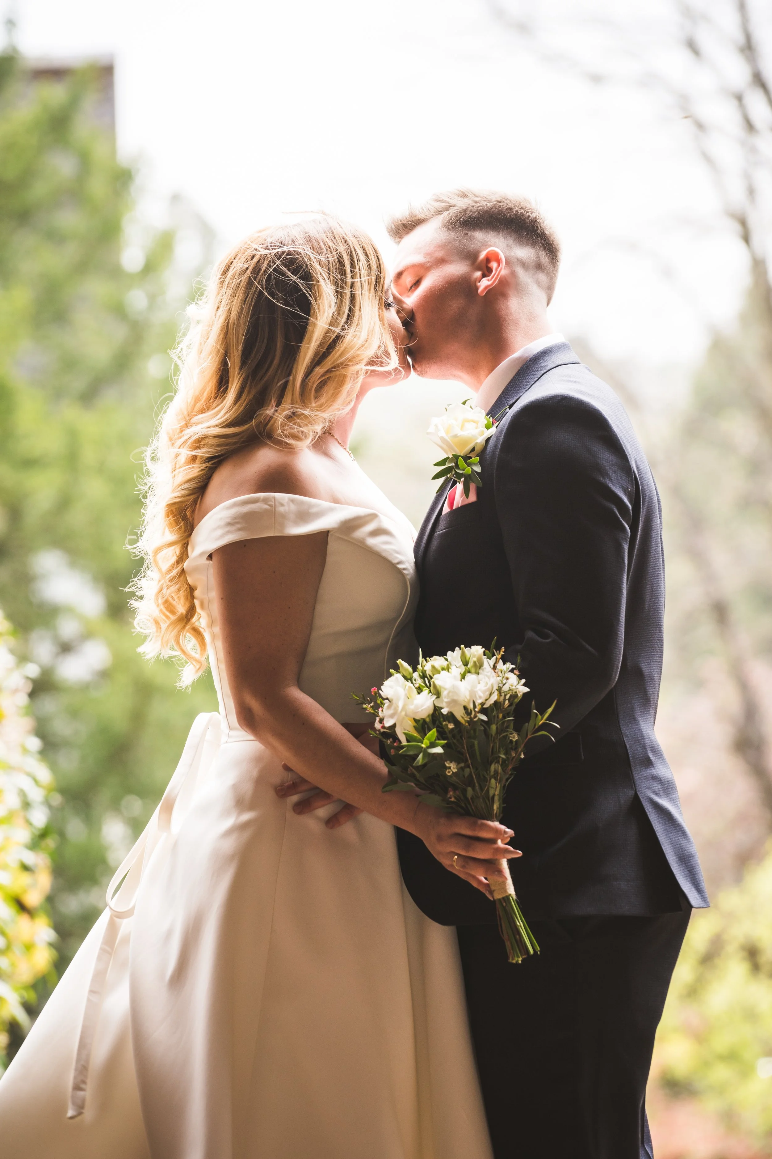 A bride and groom sharing a kiss outdoors, with the bride holding a bouquet of white flowers, and the groom wearing a dark suit with a white rose boutonniere.