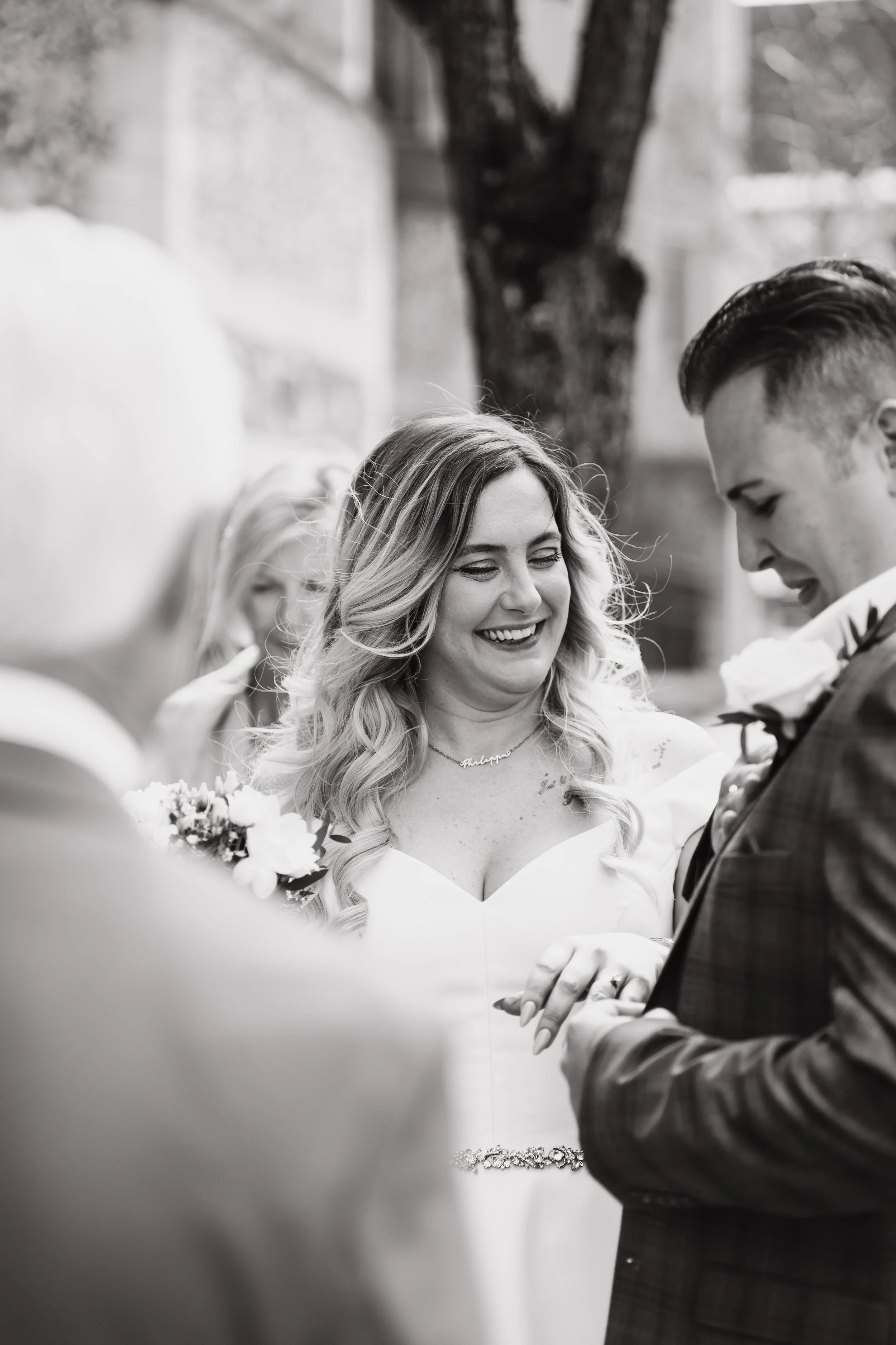 A black-and-white photo of a bride and groom during their wedding ceremony, with a woman in the foreground and a woman with glasses in the background. The bride is smiling and looking at the groom, who is holding her hand. They are outdoors near a tr