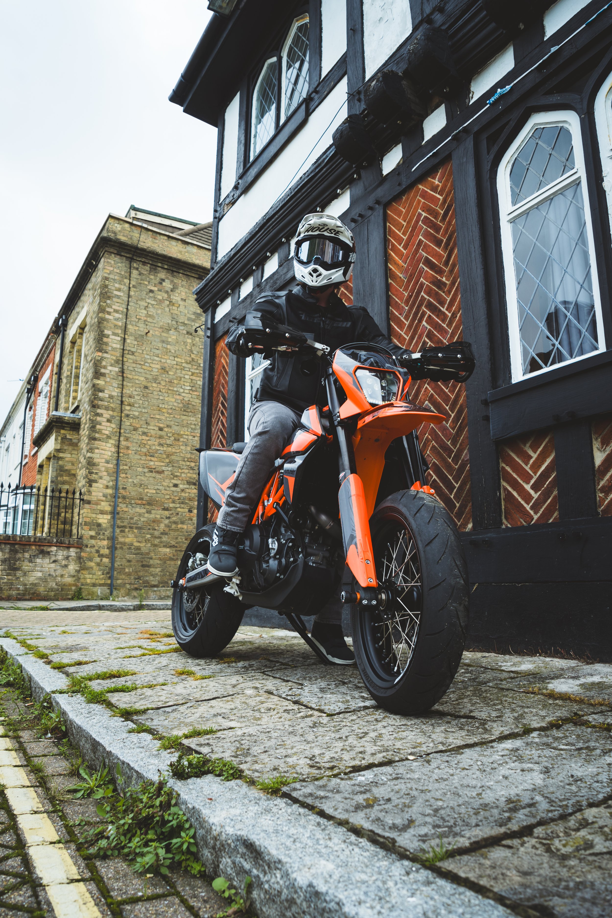 Person in motorcycle gear sitting on an orange dirt bike parked on a cobblestone sidewalk next to a black and white Tudor-style house.