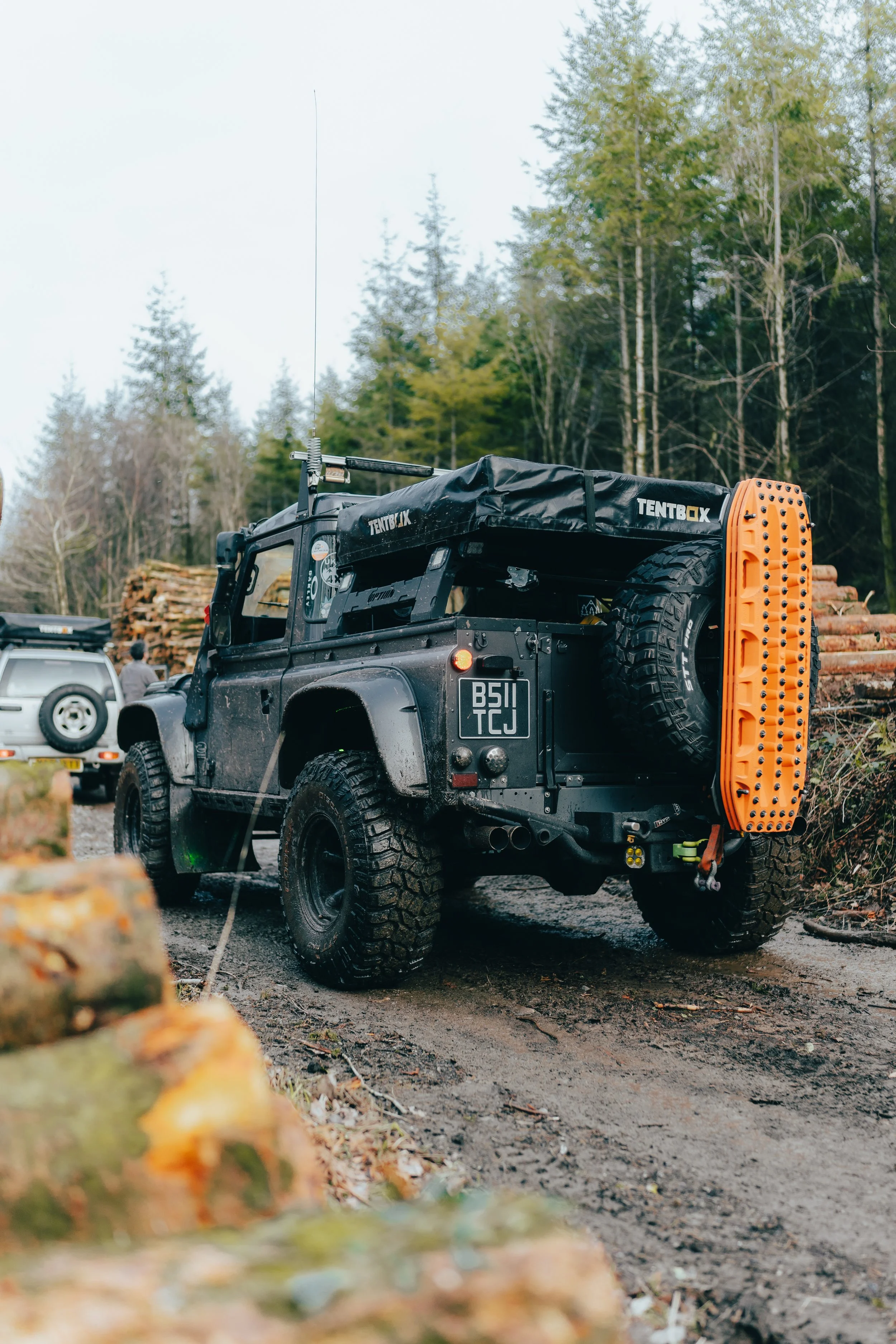 An off-road black vehicle equipped with large tires and a mounted recovery gear on the back, parked on a dirt path in a forested area with logs and other vehicles nearby.