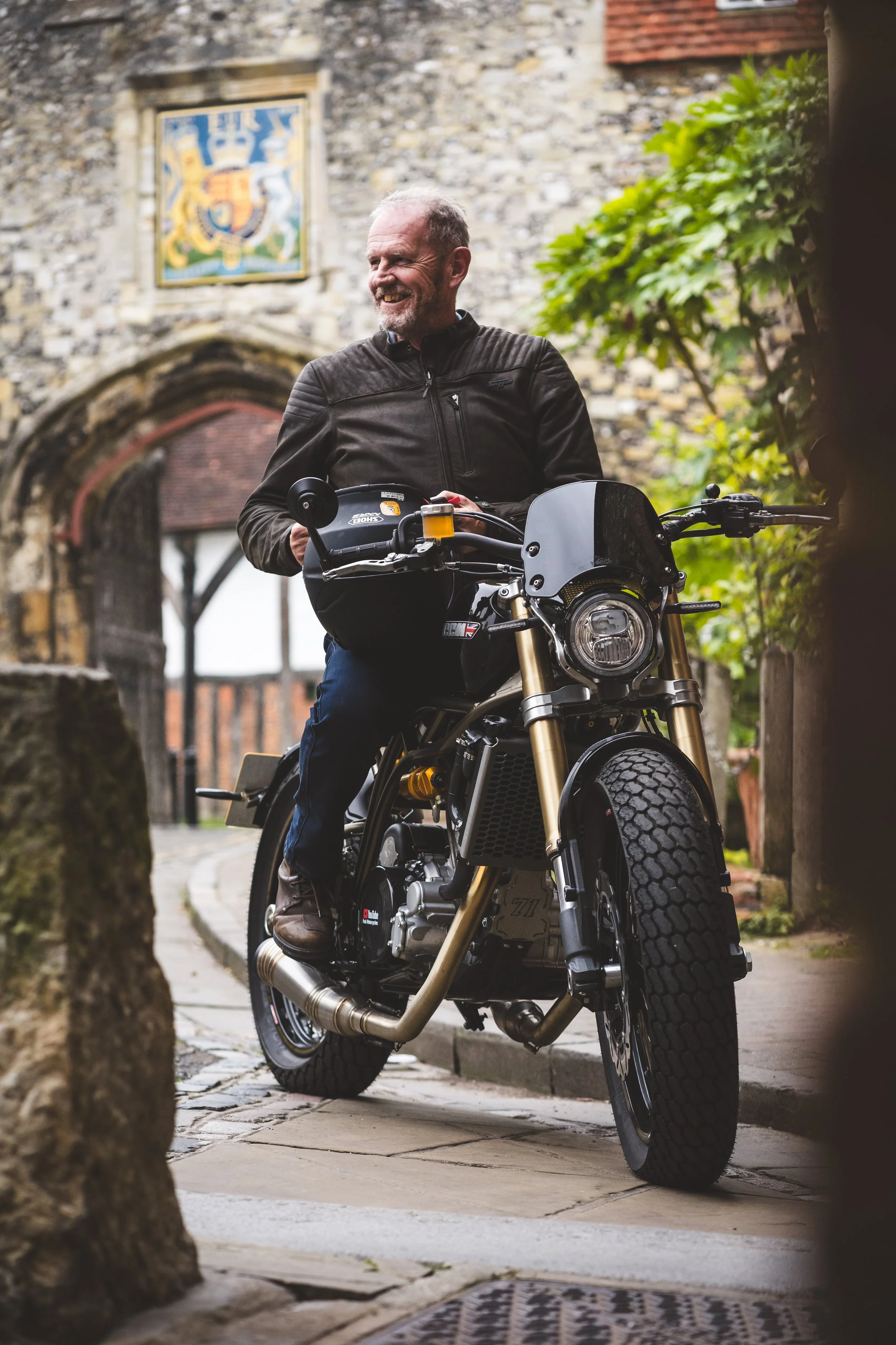 A man in a black jacket sits on a modern motorcycle with gold forks, parked on a cobblestone street near an old stone building with an arched entrance and colorful crest above it, surrounded by green foliage.