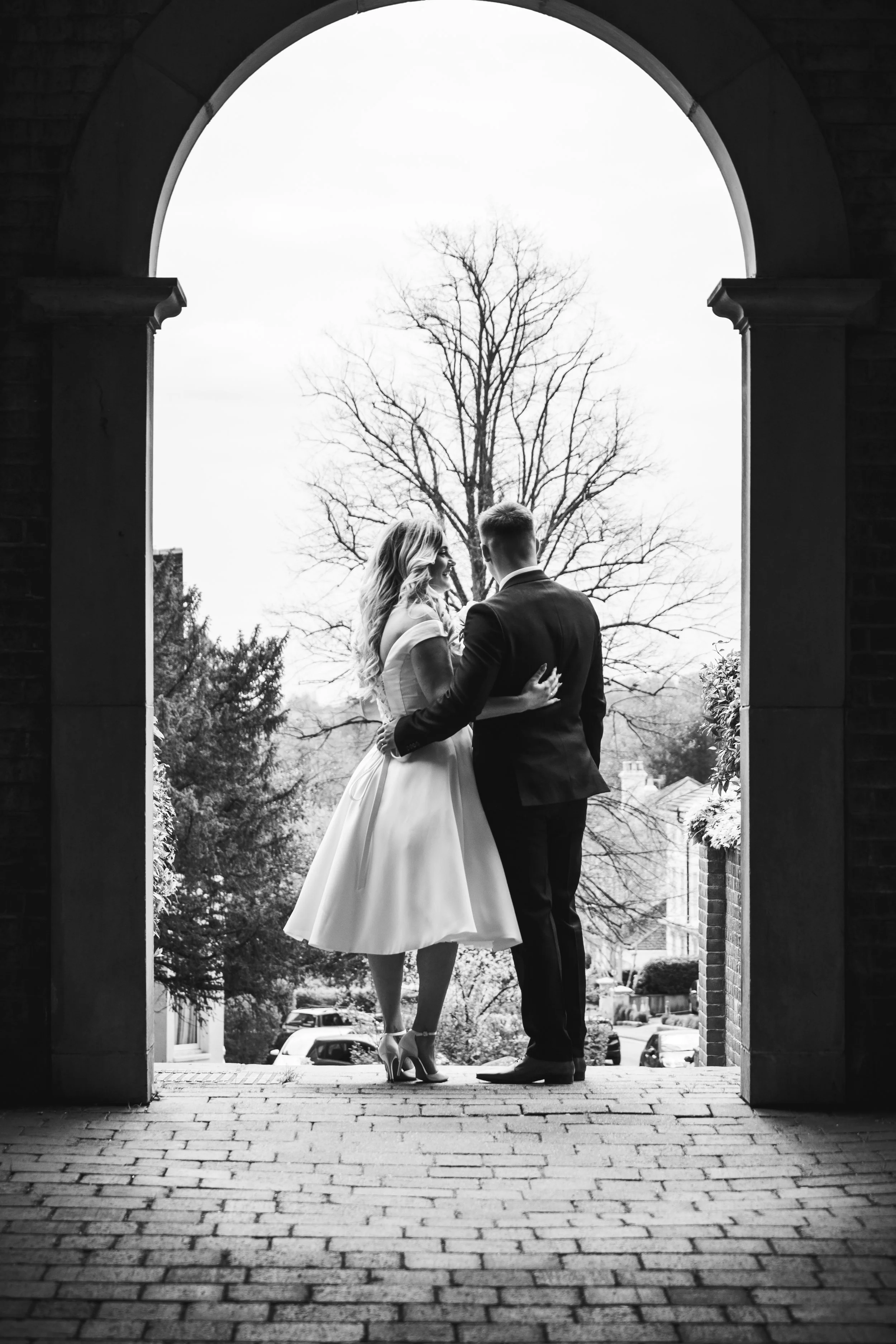 A black and white photo of a couple in wedding attire standing under an arched stone doorway, embracing each other. The woman is wearing a knee-length dress and heels, while the man is in a suit. Behind them is a large leafless tree and a cloudy sky.