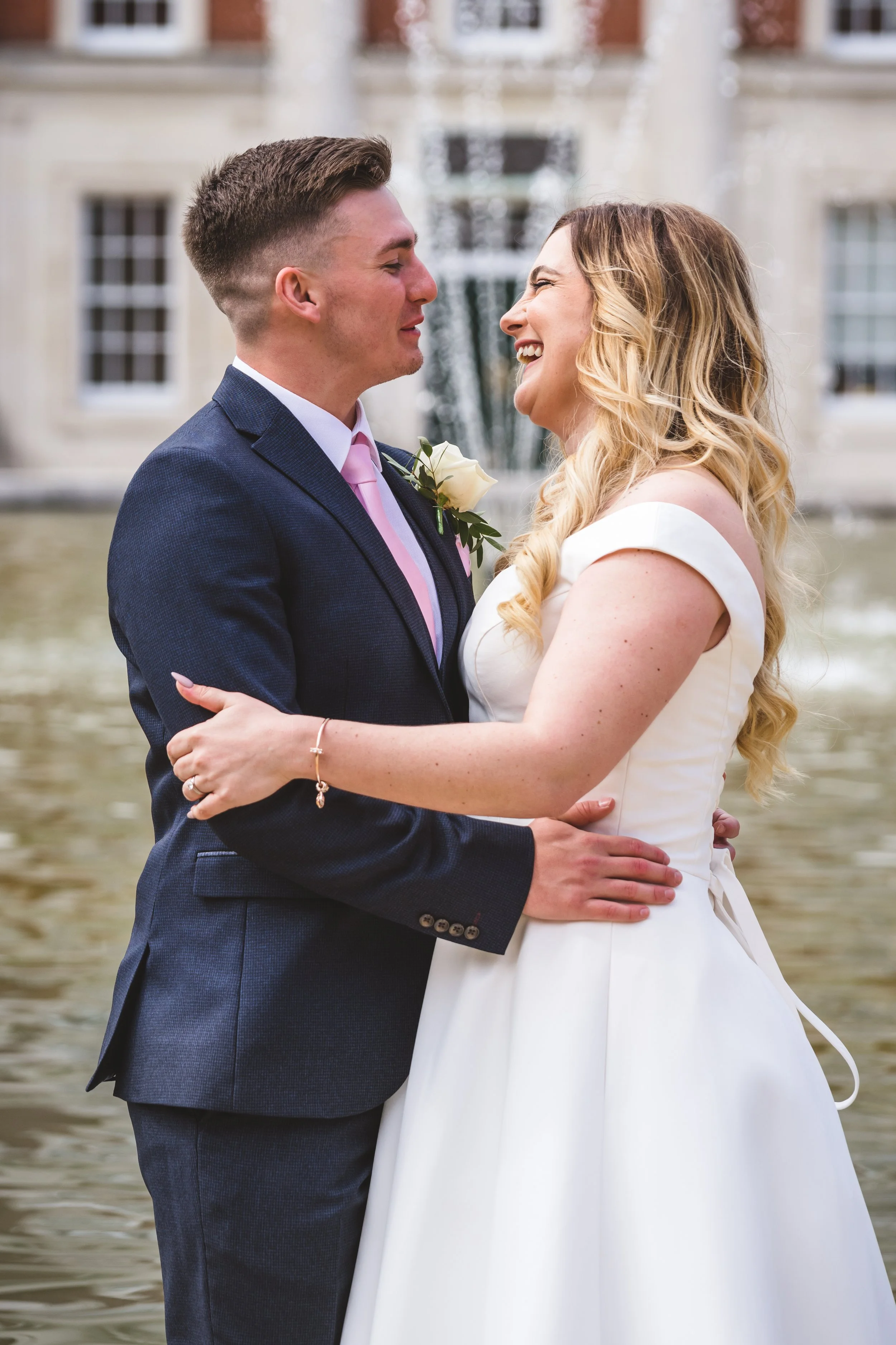 A newlywed couple dressed in wedding attire sharing a joyful moment outdoors by a fountain.