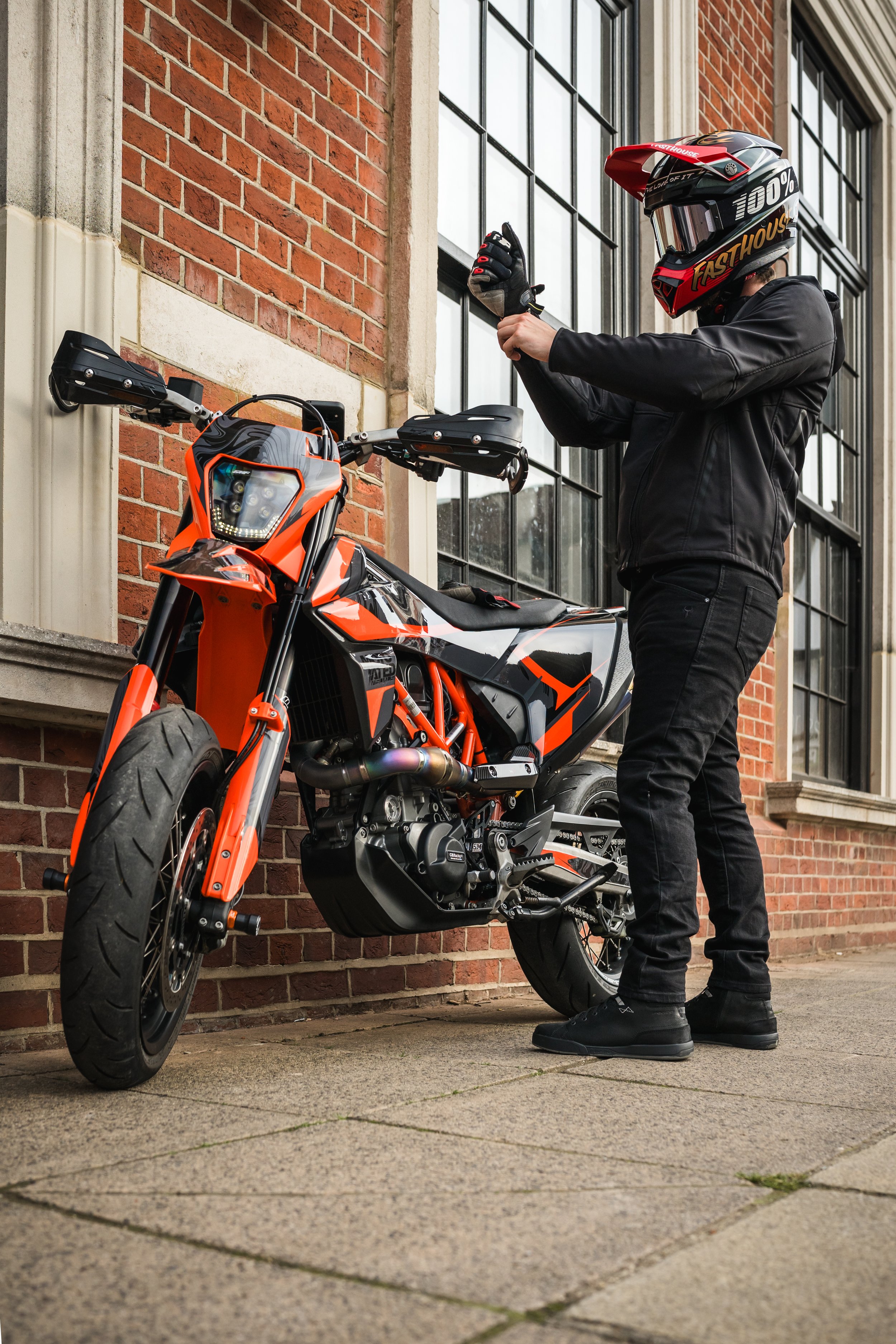 Person wearing a helmet and black jacket standing next to a black and orange motorcycle on a sidewalk.