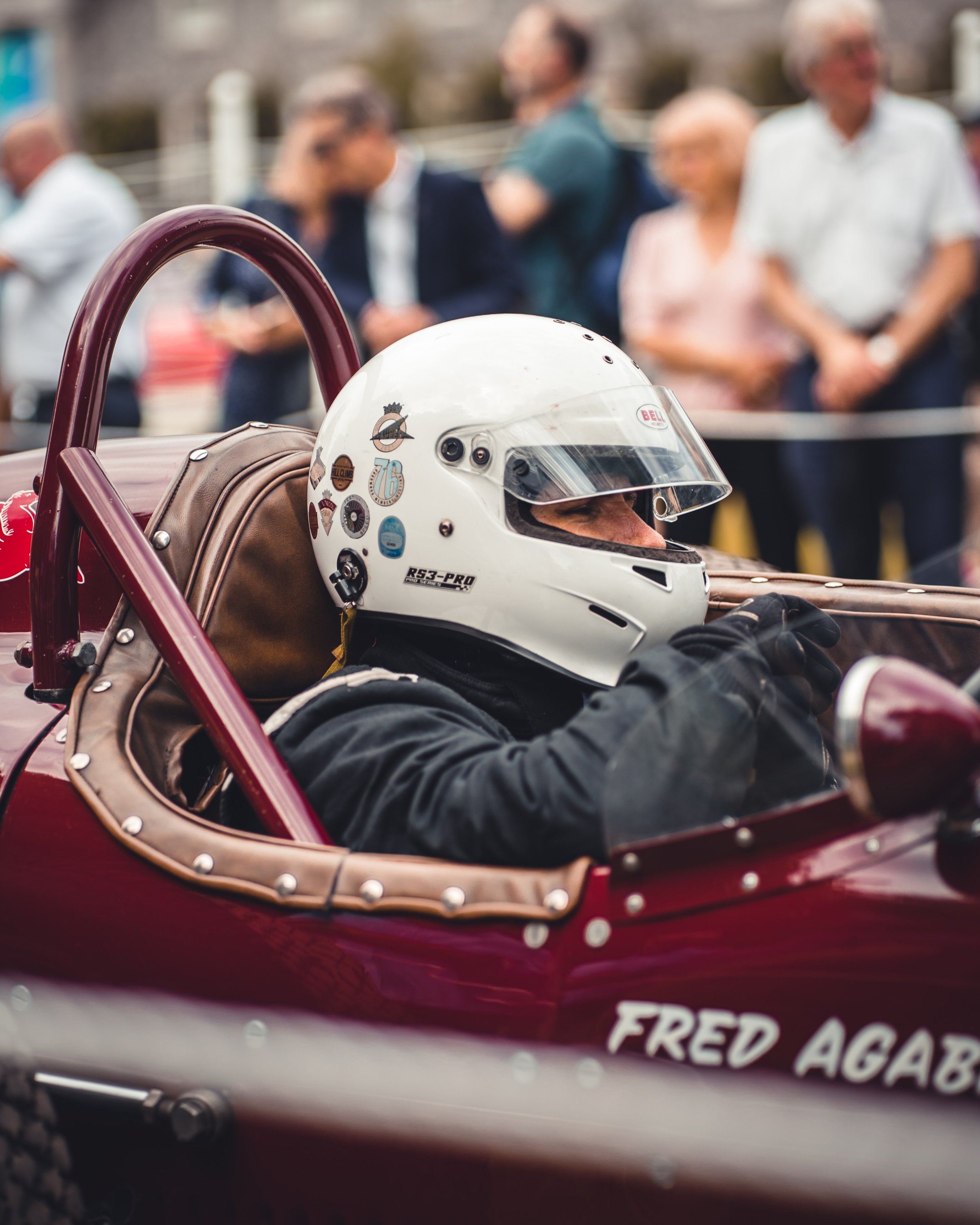 A race car driver in a white helmet and black racing suit sitting in a vintage red race car, with a group of people blurred in the background.