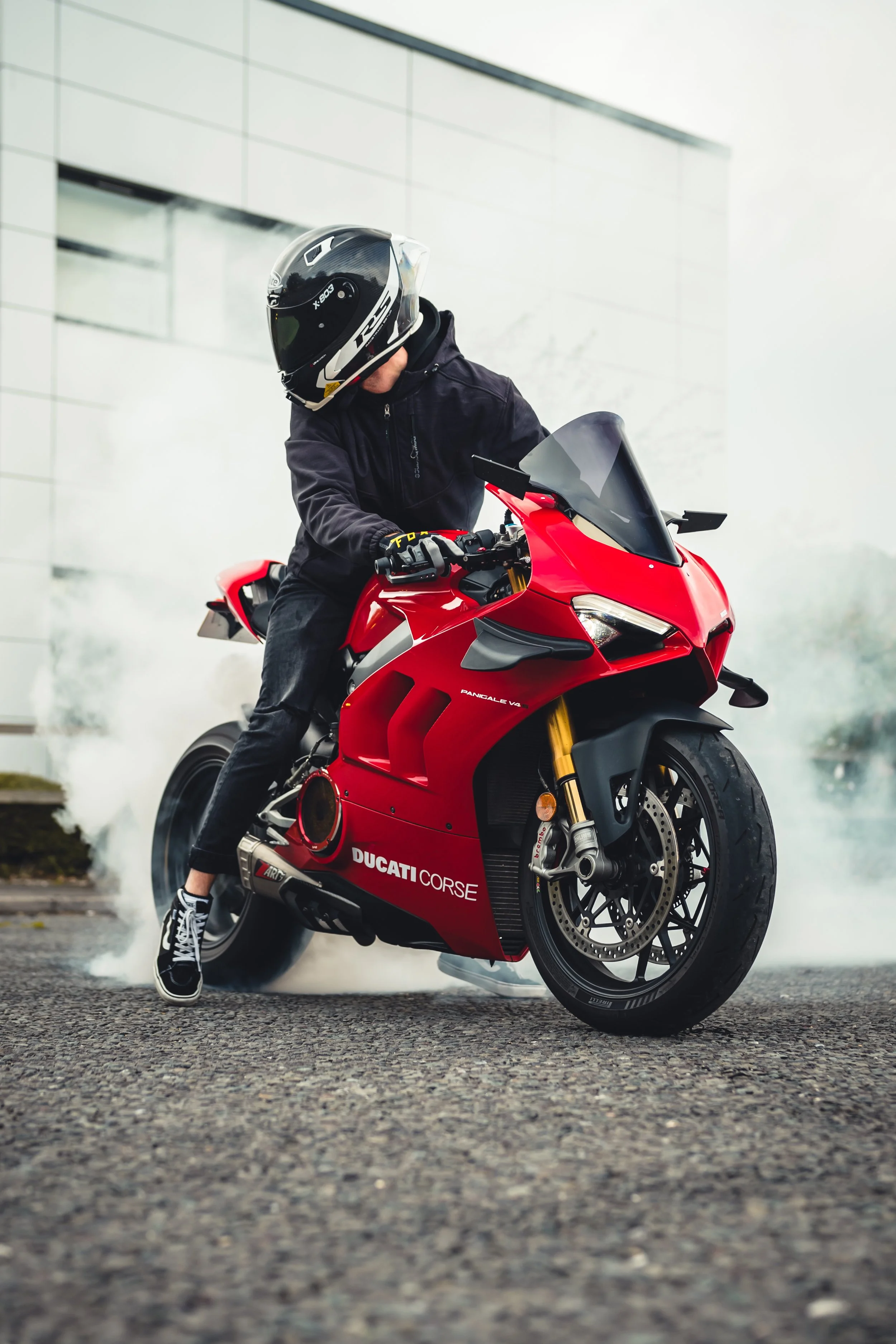 Motorcycle rider in black gear and a helmet riding a red Ducati Panigale V4 motorcycle, with smoke coming from the tires, in front of a modern building.