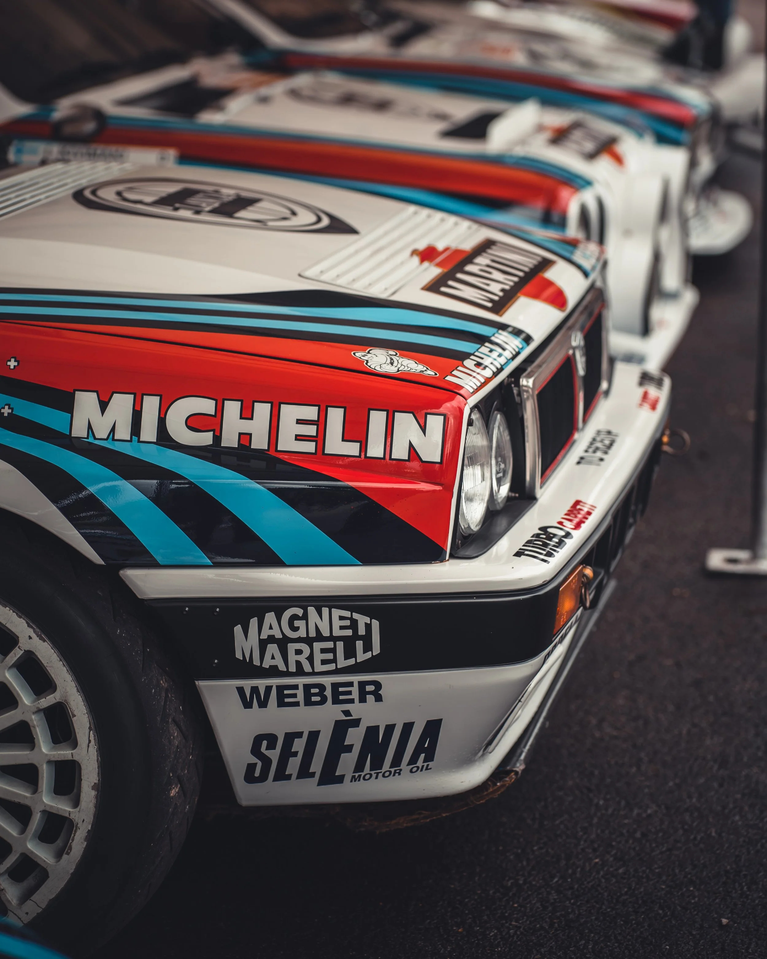 Close-up of vintage race car with Michelin sponsorship decals, showing part of the car's front and hood with blue, red, and white paint scheme, and additional sponsor logos.
