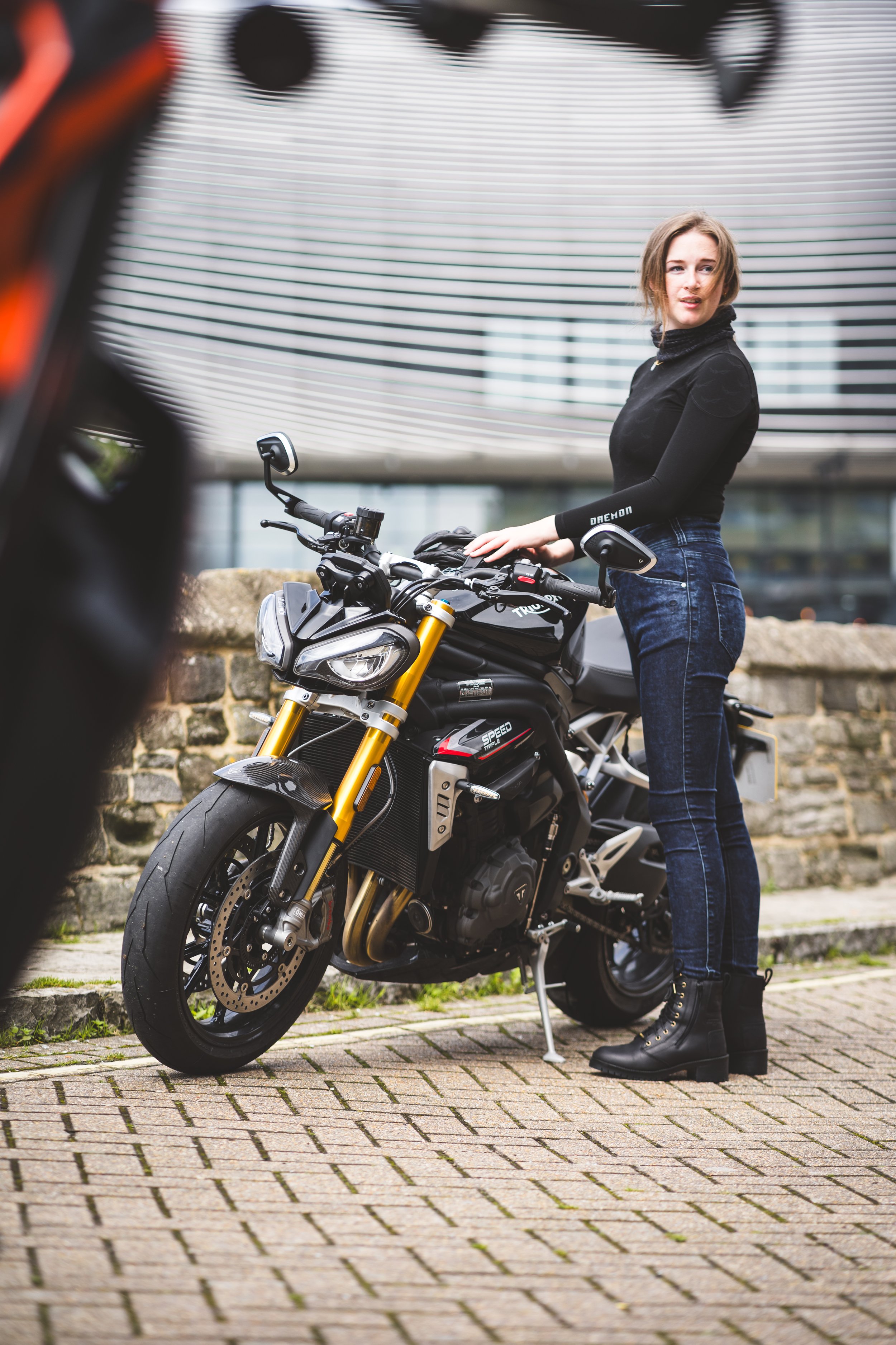 A woman standing next to a black electric motorcycle on a brick sidewalk, with modern buildings in the background.