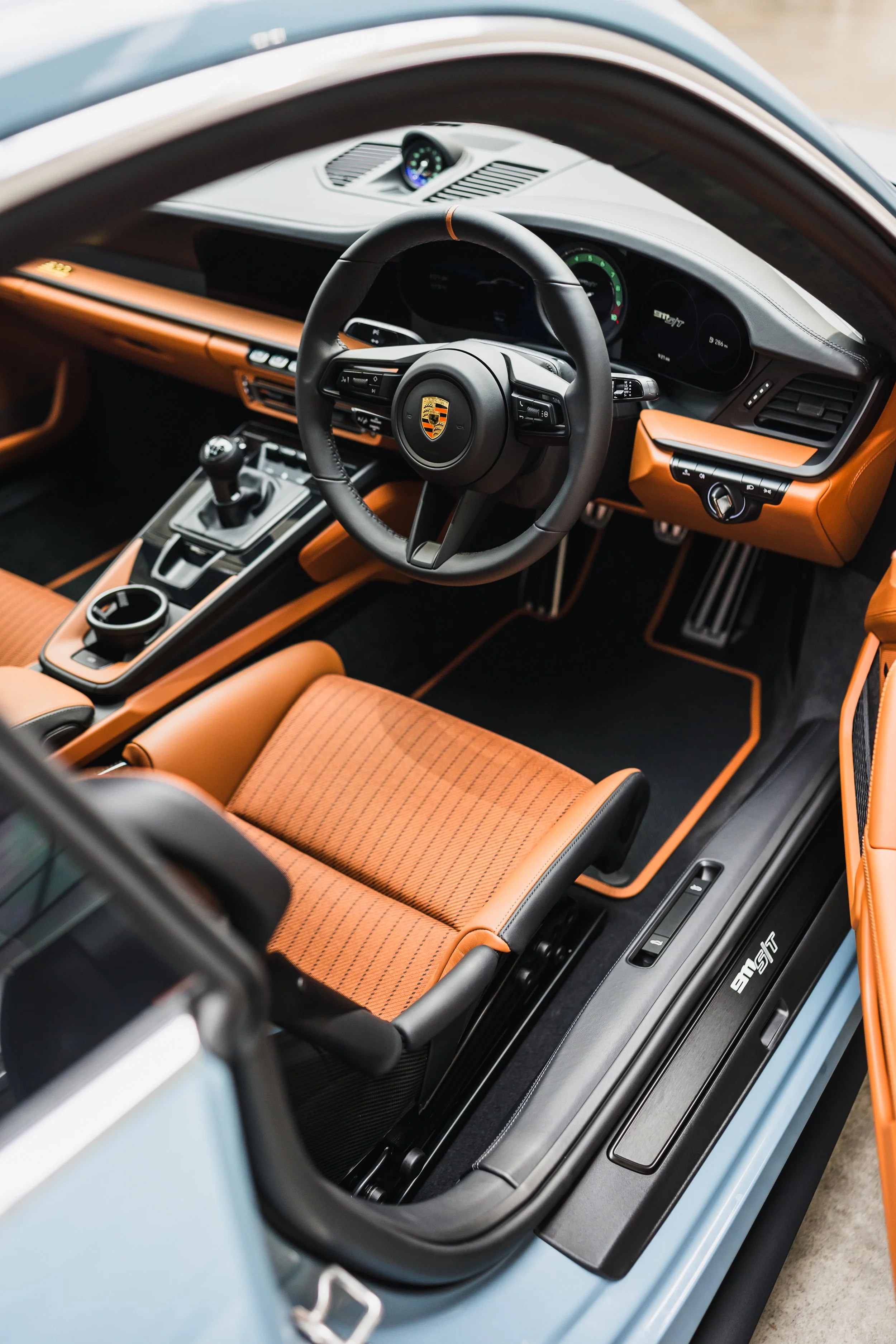 Interior view of a premium sports car with a black steering wheel featuring a Porsche logo, beige and black dashboard, gear shift, digital displays, and orange leather seats.