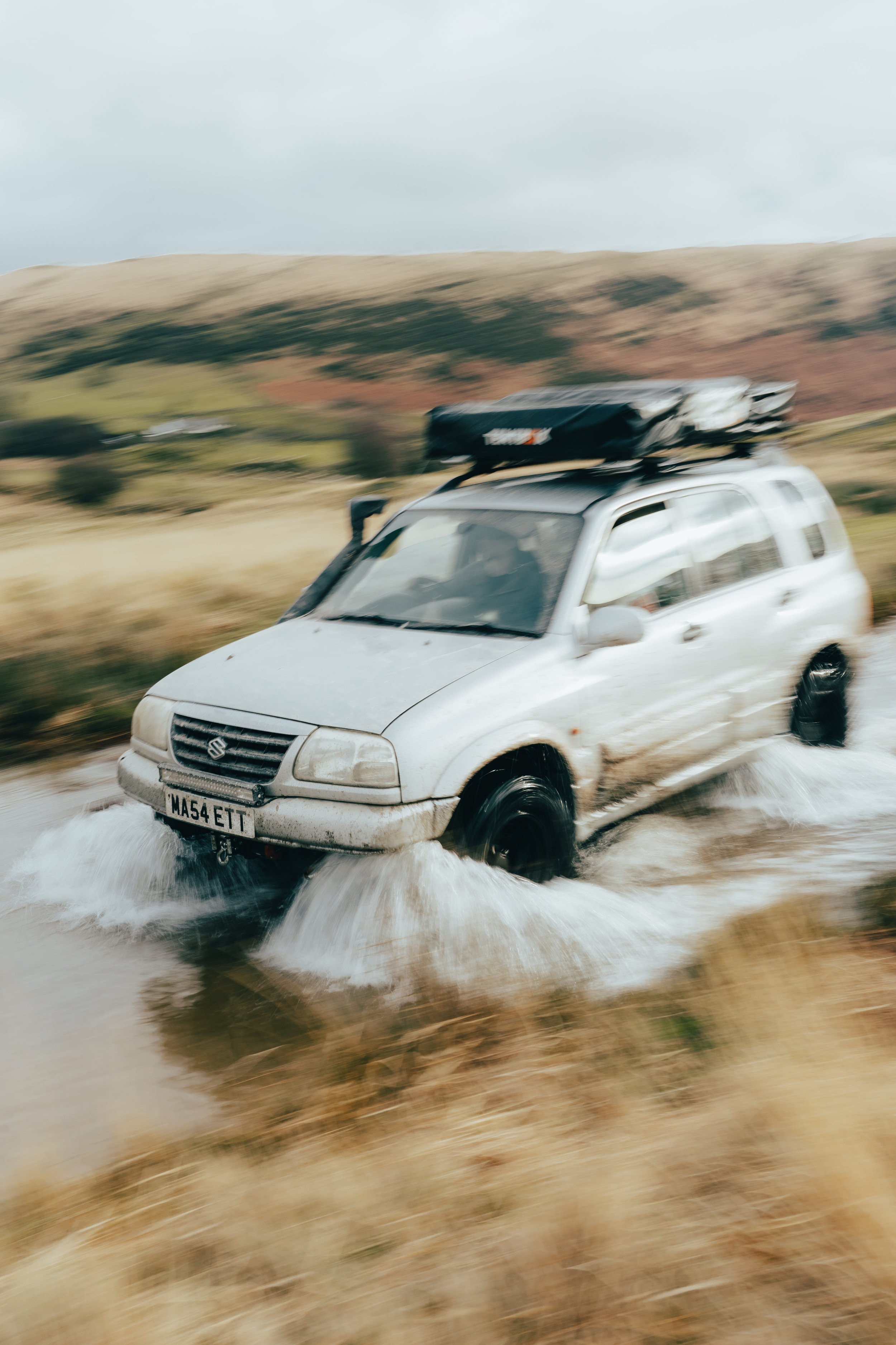 White Suzuki SUV driving through a muddy waterlogged field with hills in the background