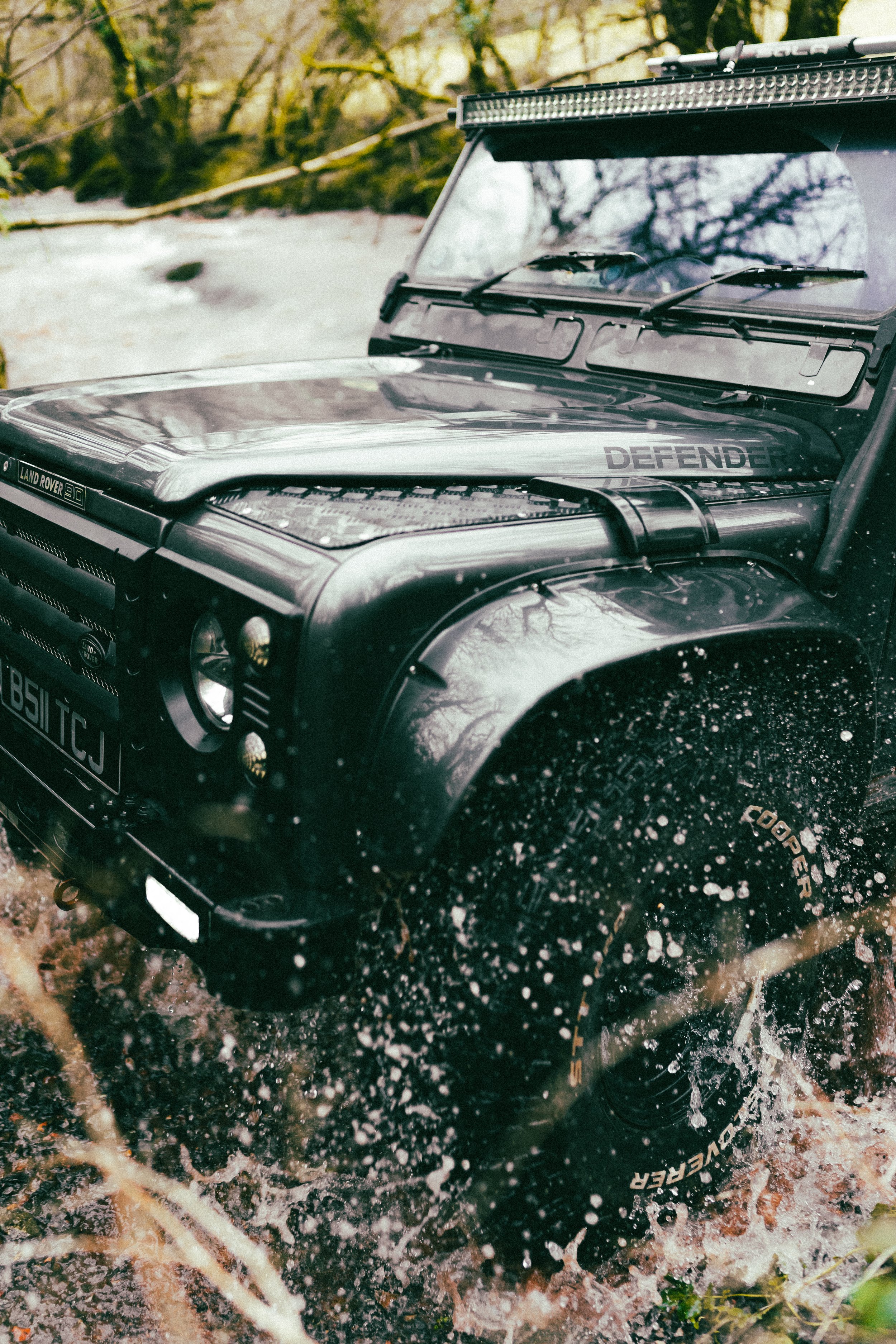 A black toy Land Rover Defender model vehicle driving through water in a natural outdoor setting.