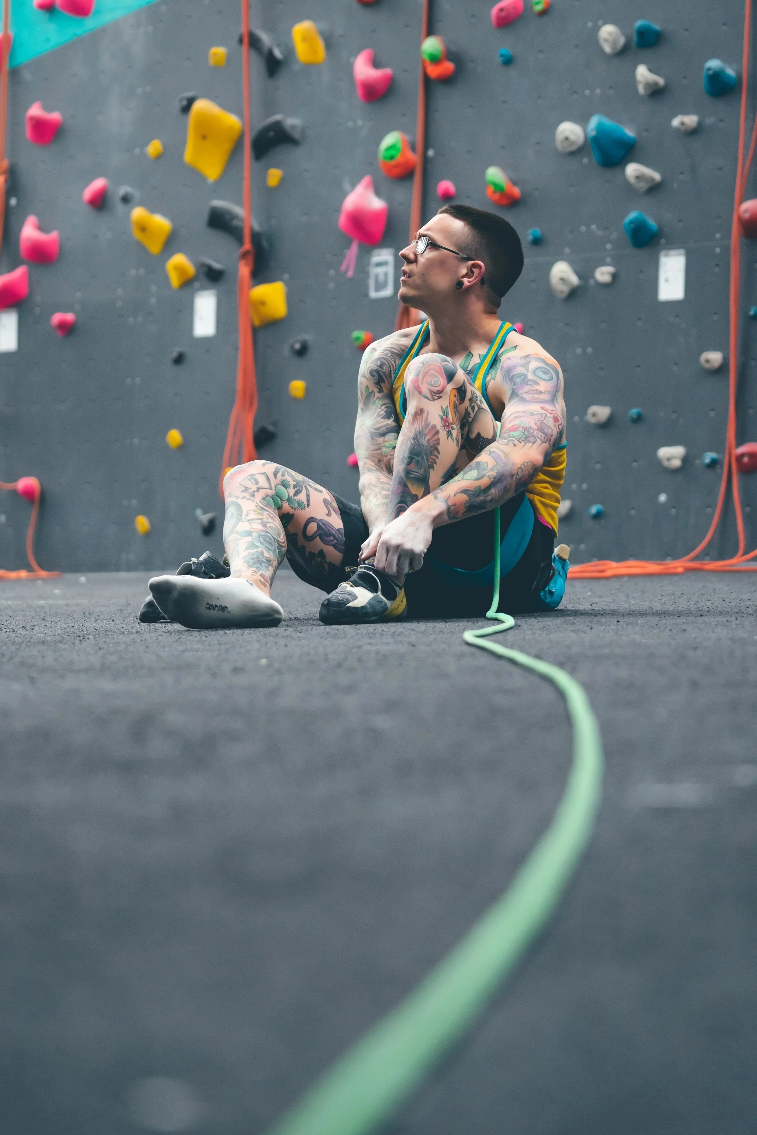 Tattooed male climber sitting on the floor at an indoor climbing gym, tying his climbing shoes, with a colorful climbing wall in the background.