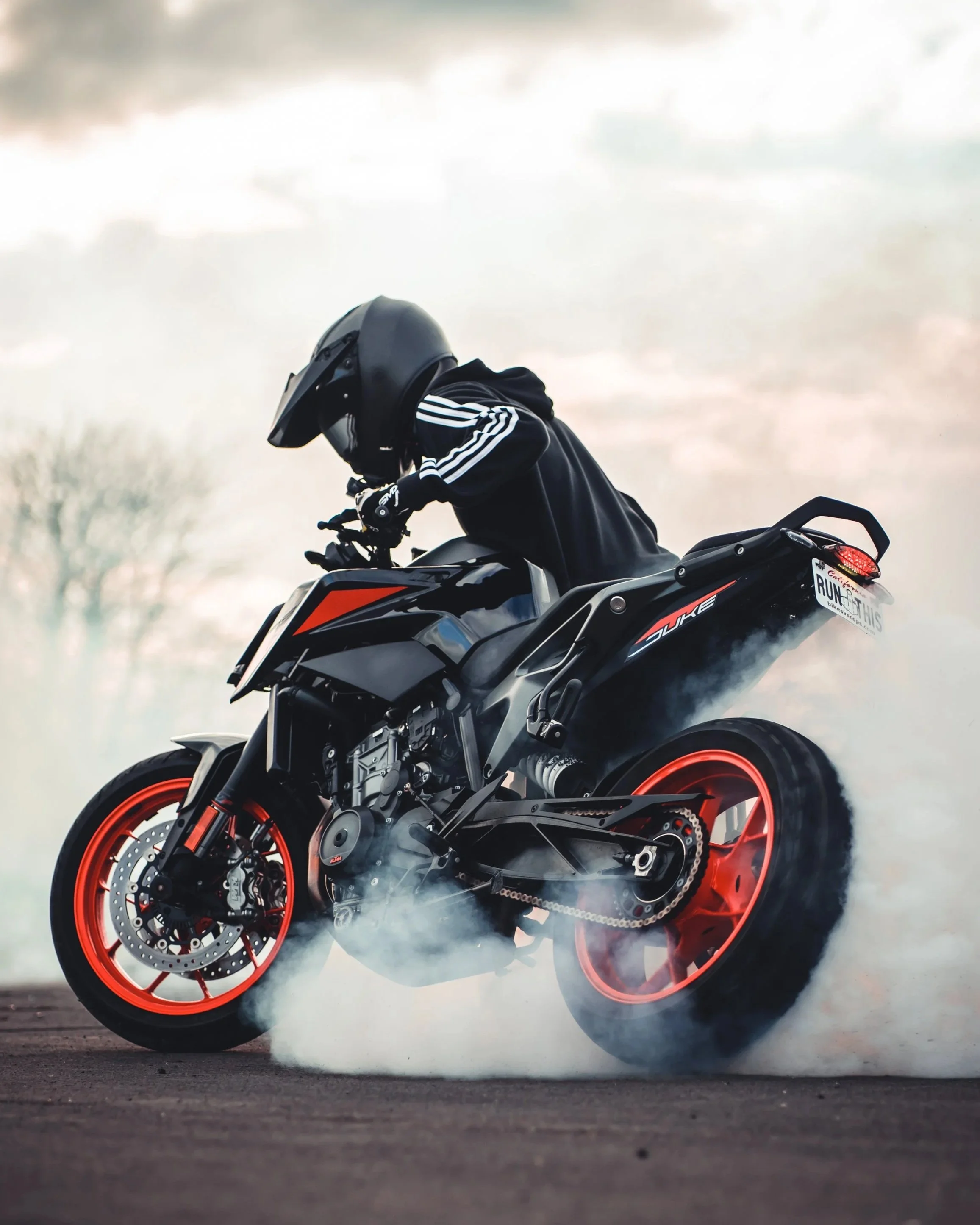A motorcyclist wearing a black helmet and black jacket performing a burnout on an orange and black sport motorcycle, with smoke billowing from the rear tire.