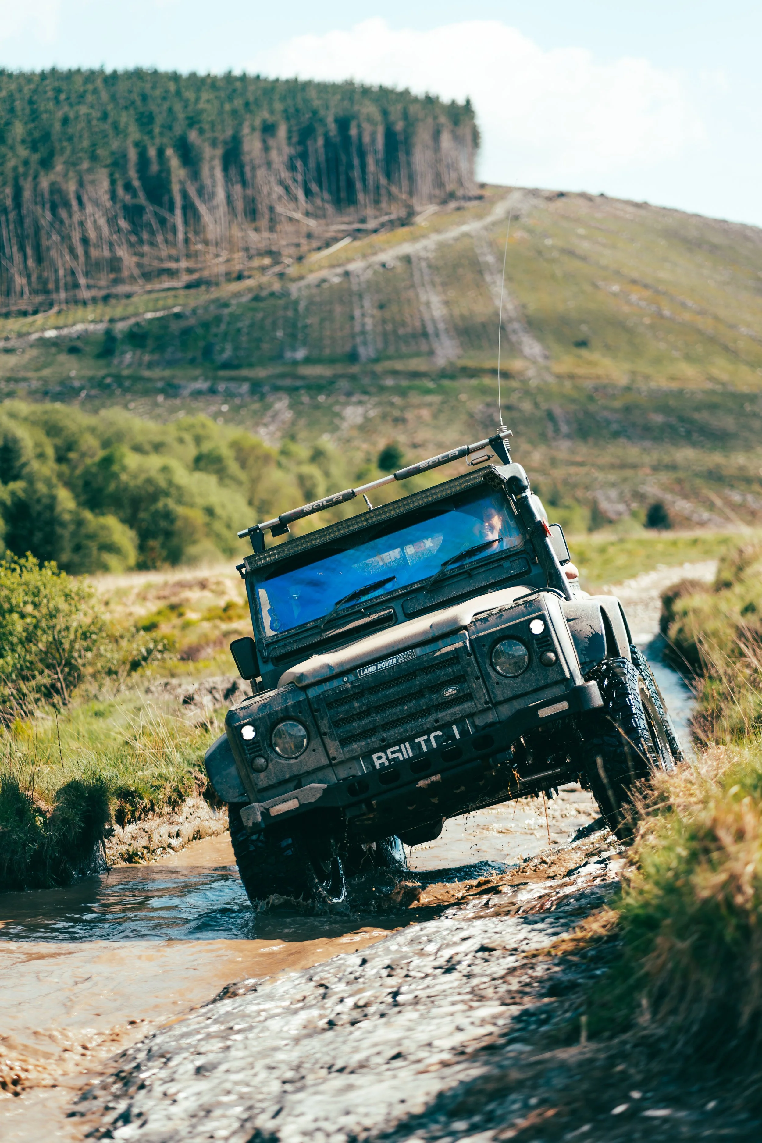A rugged Land Rover vehicle navigating a muddy water crossing on an off-road trail in a mountainous landscape with green trees and hills in the background.