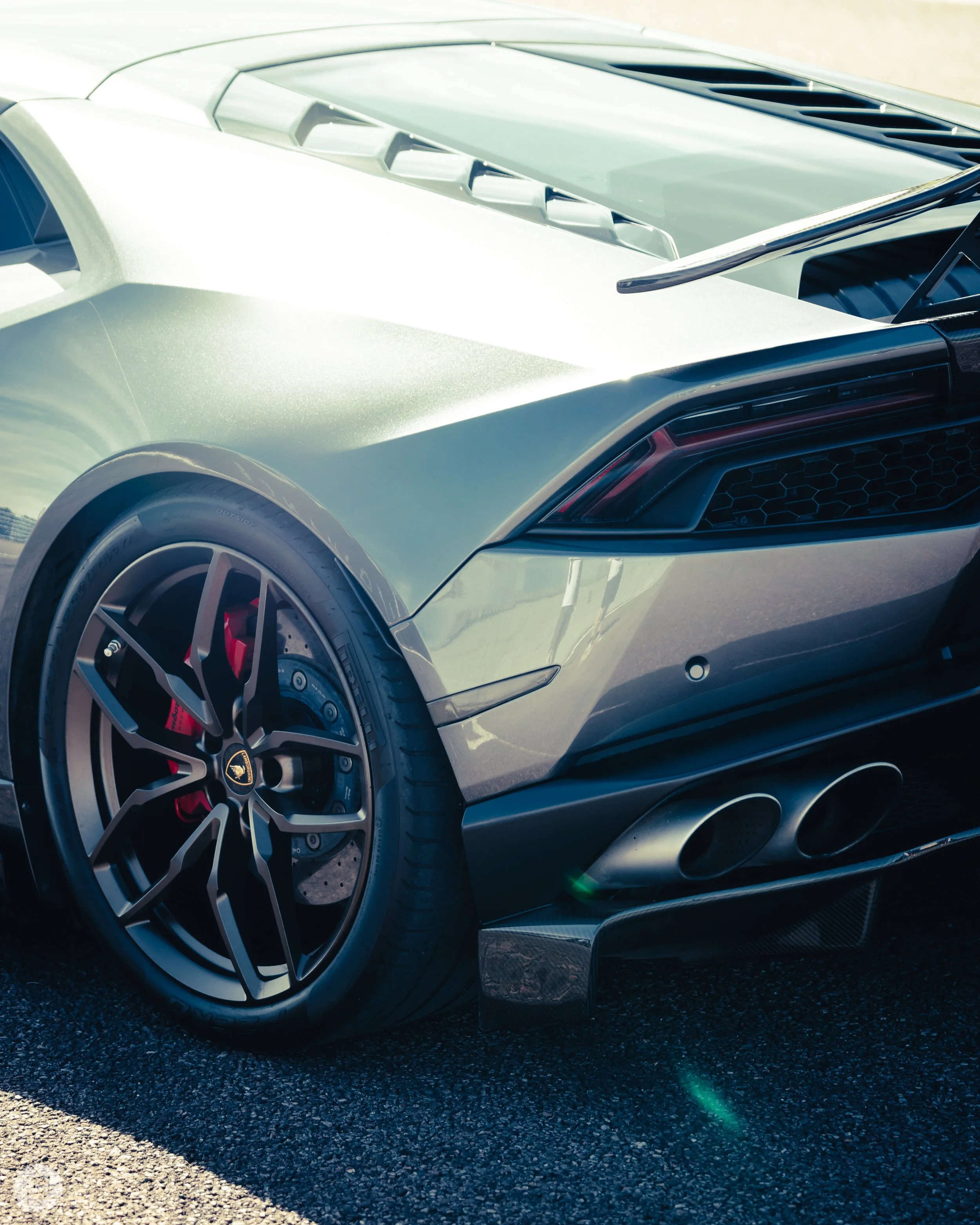 Close-up of the rear side of a silver Lamborghini sports car, showing the rear wheel with a black and silver rim, red brake calipers, and exhaust pipes at the bottom.