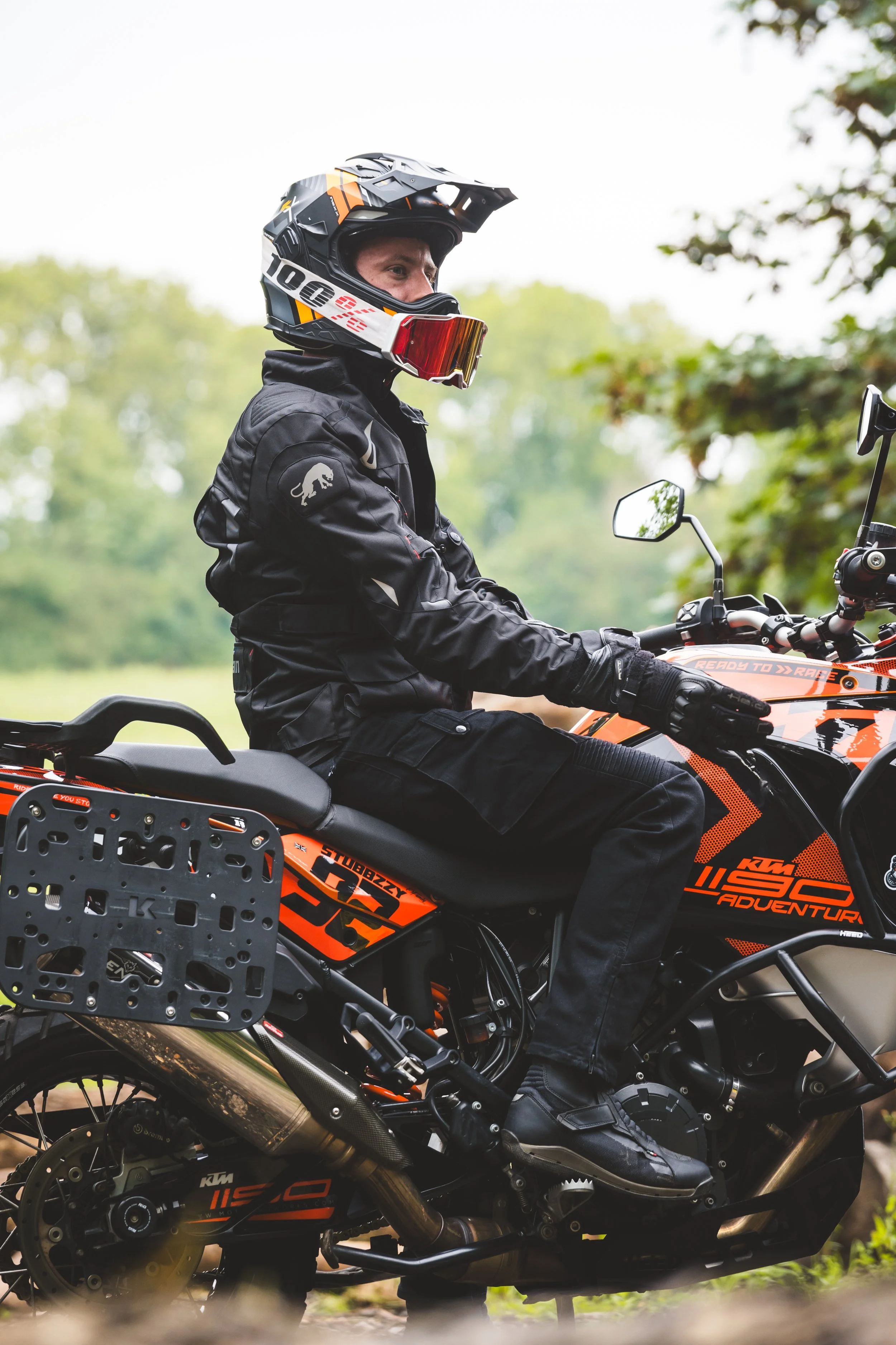A person wearing a black jacket, helmet with goggles, and black gloves sitting on an orange and black KTM 1290 adventure motorcycle outdoors.